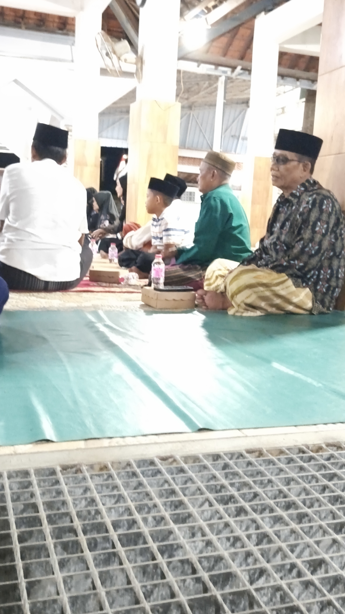 The image shows a group of men and a boy sitting on the floor, presumably in a mosque or religious building, in Dalegan, Indonesia. They are all wearing traditional Islamic attire, including peci hats. A green tarp is spread on the floor in front of them. In the background, there are wooden pillars and a tiled ceiling. There is also a metal grate visible in the foreground.
