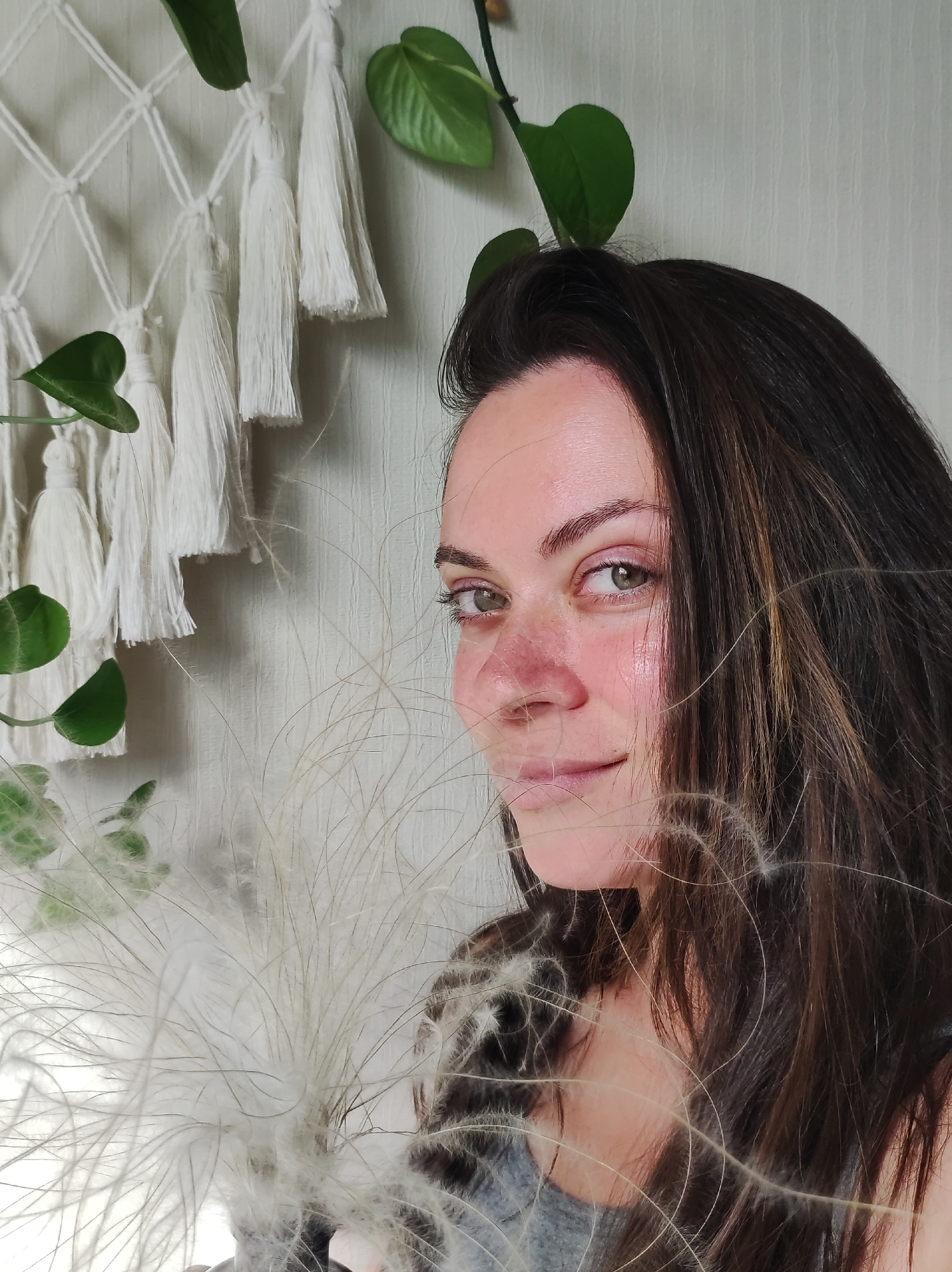 A close-up portrait captures a young woman with long, dark brown hair and light green eyes in an indoor setting in Chaikovsky, Russia. She looks directly at the camera with a soft, gentle smile. Her fair skin features visible freckles and a natural flush on her cheeks and nose.
In the foreground, a fluffy, light-colored, feathery object, resembling dried pampas grass, is positioned, partially obscuring her lower face and flowing through her hair. The background reveals a cozy interior, with a white macrame wall hanging featuring tassels prominently on the left. Green leaves from a houseplant, likely a Pothos, are intertwined with the macrame and extend into the upper parts of the frame. The wall behind her is a light, textured surface, possibly white or light grey. The lighting is soft and even, suggesting it is daytime. The overall mood of the portrait is calm and serene. No text is visible in the image.