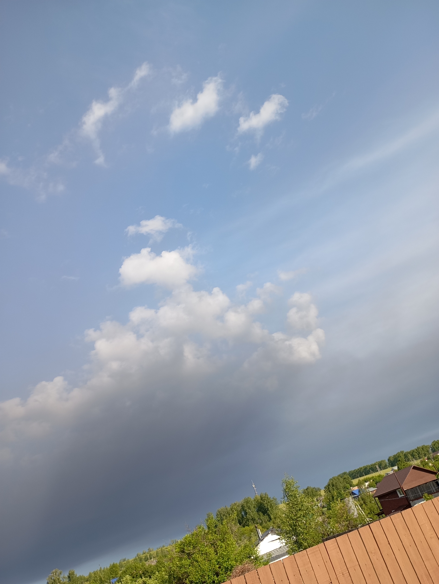 The image shows a view of the sky, likely in the city of Фоки, Russia, given the prompt's context. The sky is partially covered with clouds; some areas are clear blue, while others are filled with cumulus clouds, some of which are darker, indicating possible rain. Below the sky, there is a line of green trees and vegetation. Part of a brown wooden fence is visible in the foreground, suggesting the photo was taken from someone's property. In the background, there are hints of buildings and structures, typical of a residential area. No people or explicit actions are visible in the scene. The environment suggests a possible transition from a sunny day to a rainy or overcast one.