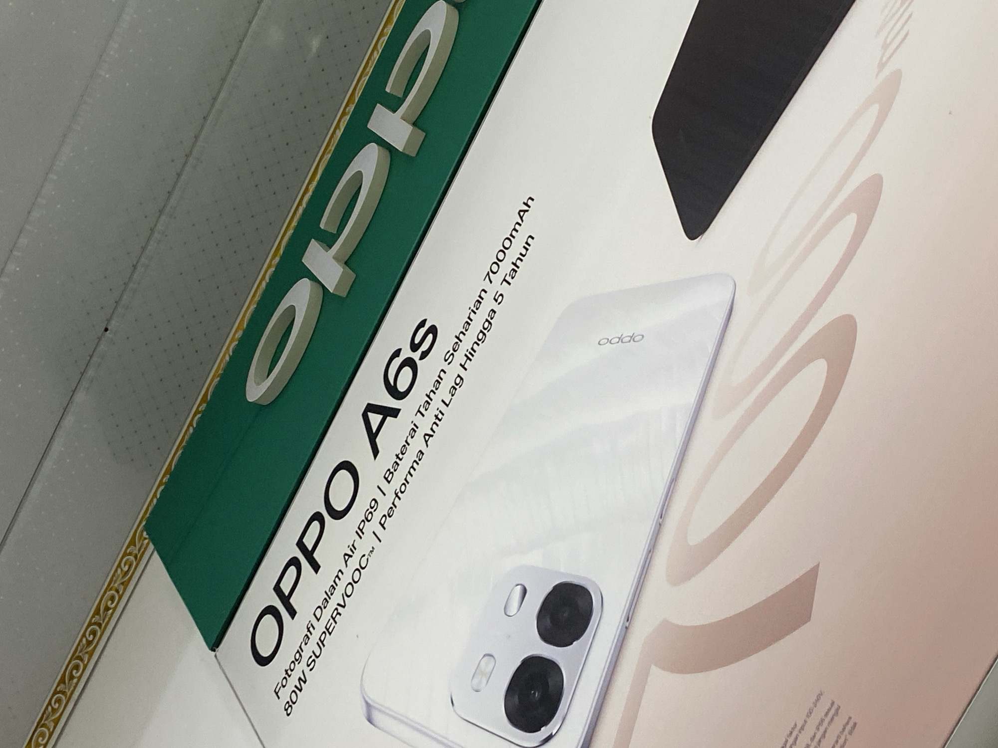 The image shows a close-up of a box for an OPPO A6s smartphone. The box is predominantly white with a green band across the top. Large white, three-dimensional lettering spells out "OPPO" above the text "A6s". Below this, there is smaller text in Indonesian that translates to "Photography in IP69 Water | 7000mAh Continuous Battery | 80W SUPERVOOC™ | Performs Anti-Lag for 5 Years". A white smartphone with a dual-camera module is positioned on the box, partially obscuring the text. The background appears to be a ceiling with a patterned border. The overall lighting suggests an indoor setting, possibly a retail display or a product advertisement. There are no people or specific landmarks visible in the image, so it is impossible to determine its location in Ta', Indonesia.