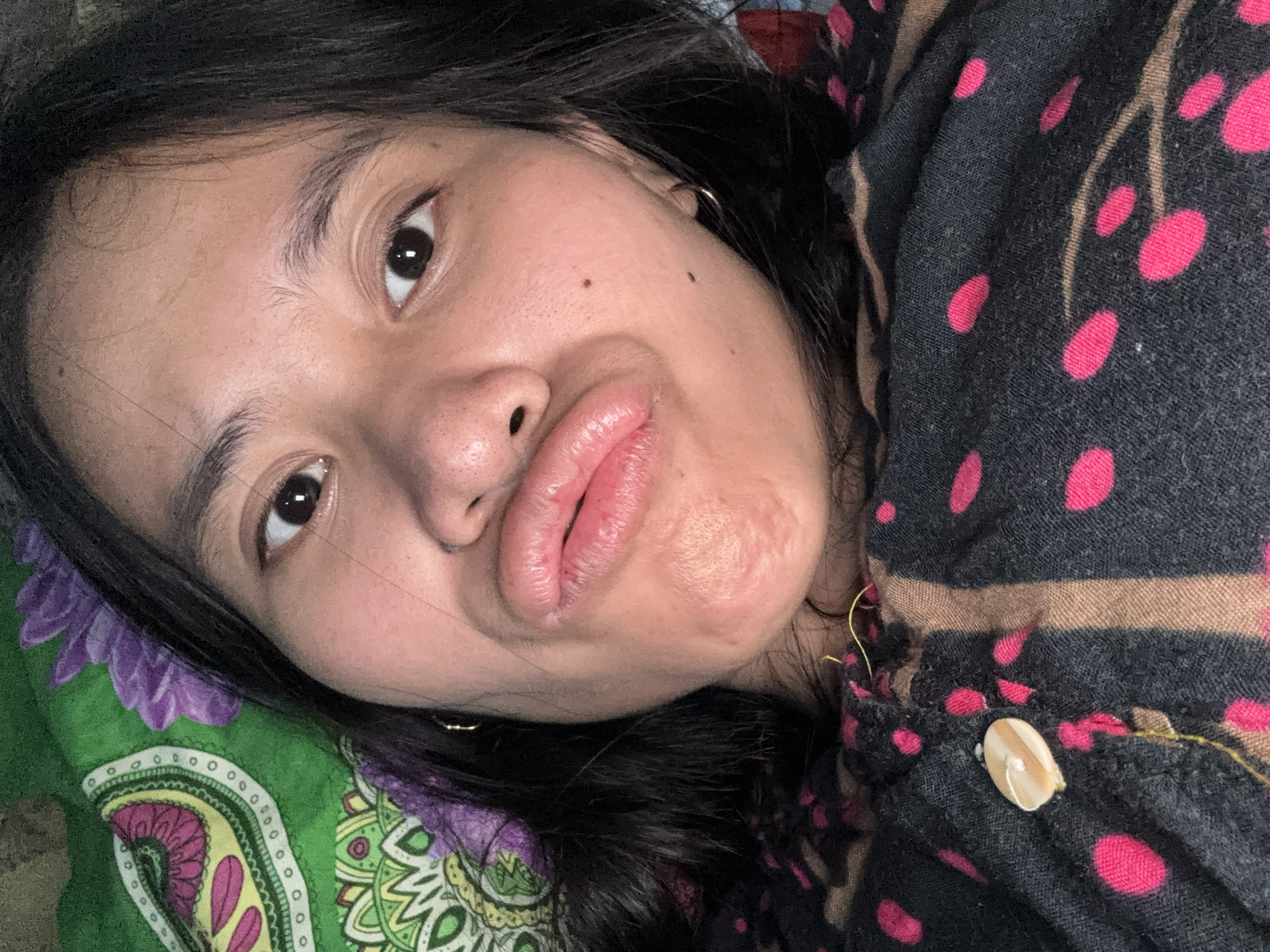 This is a selfie of a young woman in East Jakarta, Indonesia. She is lying down, looking at the camera with her lips pursed. She is wearing a dark-colored shirt with pink polka dots and brown stripes. A button on the shirt is visible. Behind her, a green fabric with a floral pattern is visible. She has dark hair and is wearing small hoop earrings. The lighting suggests indoor conditions.