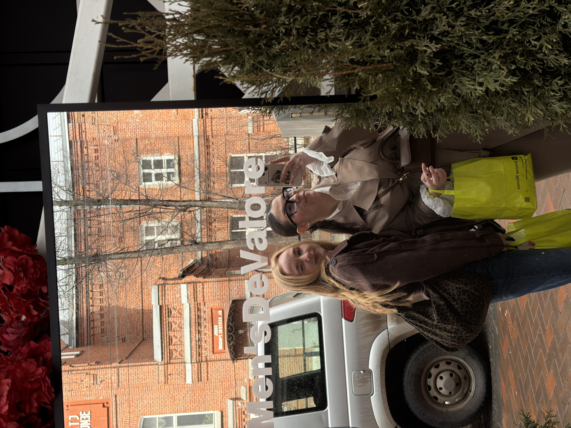 Two women are standing next to a silver van. One woman is holding a phone and taking a picture. The women are in Perm, Russia, as reflected in a mirror behind them. A building made of red brick is reflected in the mirror. There is a sign for Men's De'Vator in the reflection. One woman is wearing a brown jacket and blue jeans. The other woman is wearing a beige coat, has a yellow bag, and wears black-framed glasses. A coniferous tree is near the top of the image. Red flowers are near the lower left of the image.