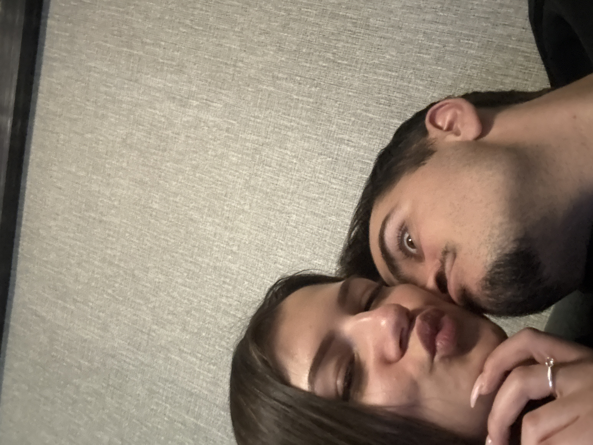 A man and a woman are close together, possibly lying down indoors, as indicated by the neutral-colored wall in the background. The woman has dark hair and is making a kissing face with her eyes closed. A ring is visible on her finger. The man, with dark hair and a beard, is positioned close to the woman's face. The location in Sofia, Bulgaria, is unknown from this image.