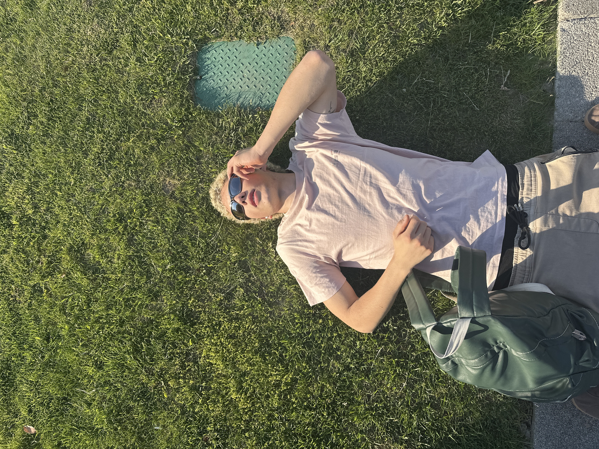 An overhead shot captures a young person relaxing on their back on a patch of vibrant green grass, bathed in bright sunlight. The scene is likely set outdoors in a park or public green space in Novi Sad, Serbia.
The person has short, light-colored (possibly bleached blonde) curly hair and is wearing dark sunglasses, a light pink short-sleeved t-shirt with a faint, indiscernible graphic or text on the front, and light beige cargo-style pants. One arm is bent, with the hand near their forehead, while the other rests on their chest area. Their posture suggests a moment of leisure and comfort.
To the upper left of the person's head, a dark green rectangular utility cover with a textured diamond plate pattern is embedded in the grass. Near their feet, to the right, a dark green duffel bag or large purse with light-colored handles rests on the grass. The right edge of the frame reveals a paved area, possibly a sidewalk or path, with the toe of a dark brown shoe visible on it.
The harsh, defined shadows cast by the person, the utility cover, and the edge of the paved ground indicate strong, direct sunlight, suggesting a clear day, likely during midday or early afternoon. The overall mood is relaxed and peaceful. No clearly legible text is visible on any objects in the image.