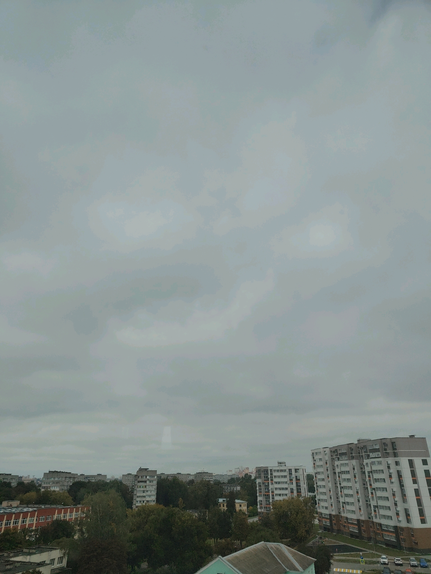 An overcast, daytime aerial view depicts the city of Homyel, Belarus. In the foreground, several multi-story apartment buildings are visible, with one building in the lower left featuring a red brick facade. The middle ground is filled with dense green foliage from trees. Further in the distance, more apartment buildings and some industrial structures are spread across the landscape. The sky is uniformly gray with clouds. No people are discernible in the image.