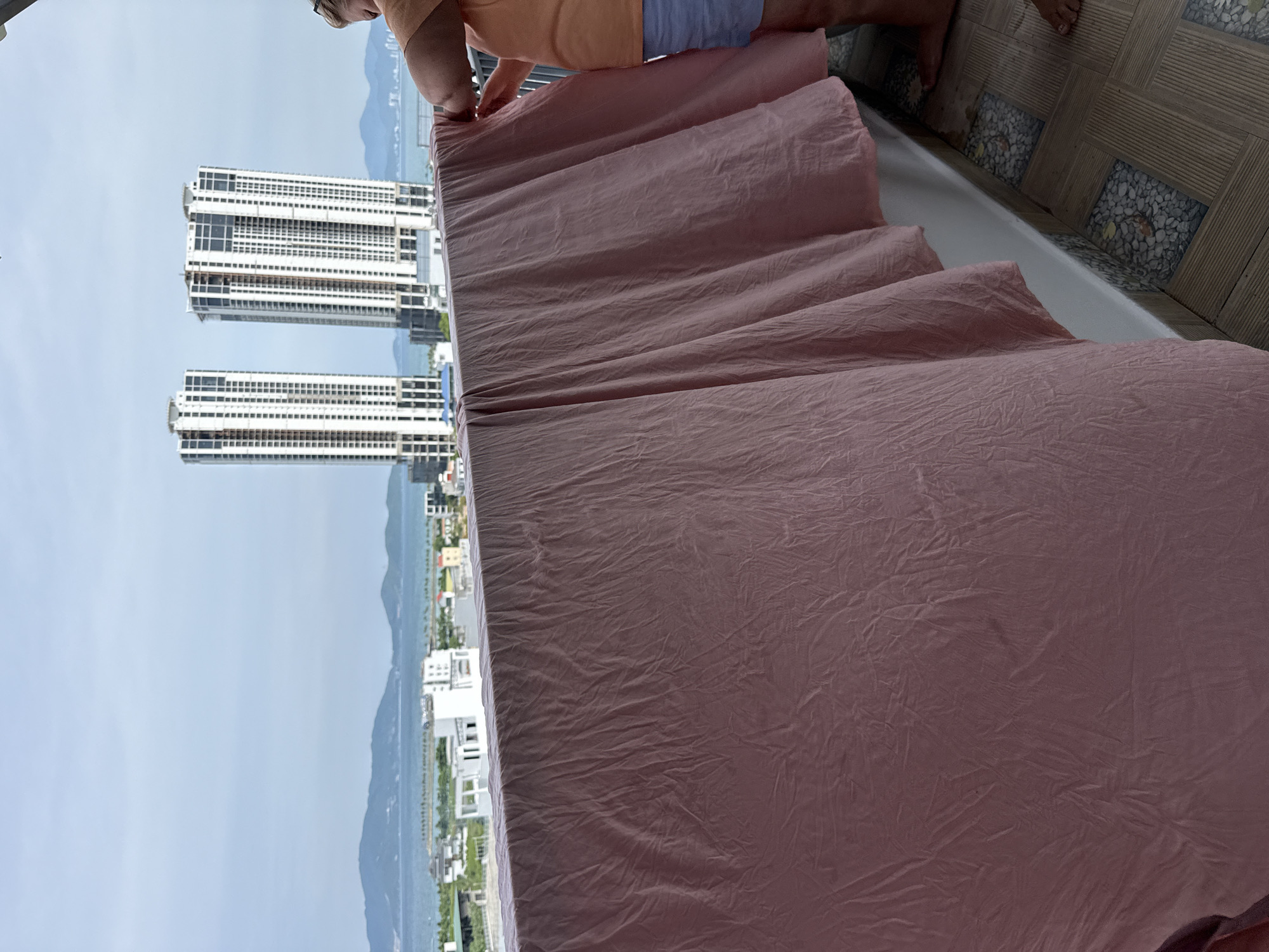 An individual is standing on a balcony in Bac Nha Trang Ward, Vietnam, hanging a pink sheet over a railing. The sheet is draped downwards, creating a large pink curtain. In the background, two tall, white skyscrapers dominate the skyline, with a city and a body of water visible beyond them. The sky is overcast. The balcony floor is tiled with a mosaic of pebbles and wood-grain patterns. The person is wearing an orange t-shirt and blue shorts.
