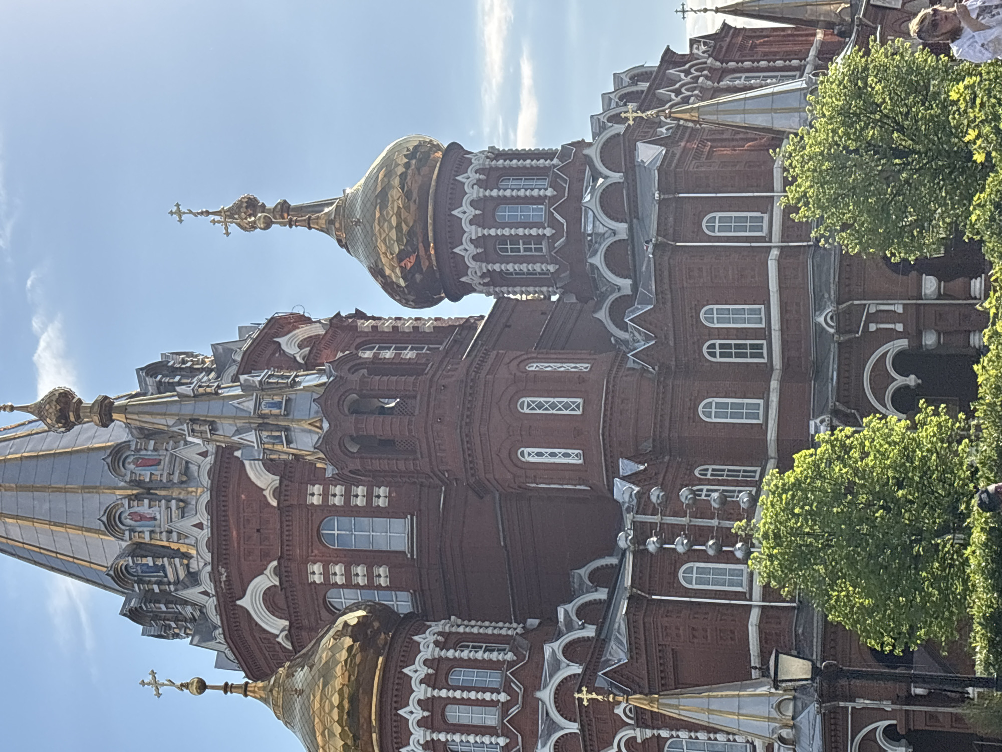The image shows a view of the architectural details of St. Michael's Cathedral in Izhevsk, Russia. The cathedral is primarily constructed of reddish-brown brick, adorned with intricate white decorative elements, numerous windows, and golden domes topped with Orthodox crosses. The domes' golden surfaces reflect the sunlight, adding to the visual richness of the scene. Some green foliage from nearby trees is visible in the bottom right corner, partially obscuring the cathedral's facade. The sky in the background is light blue with some wispy clouds.