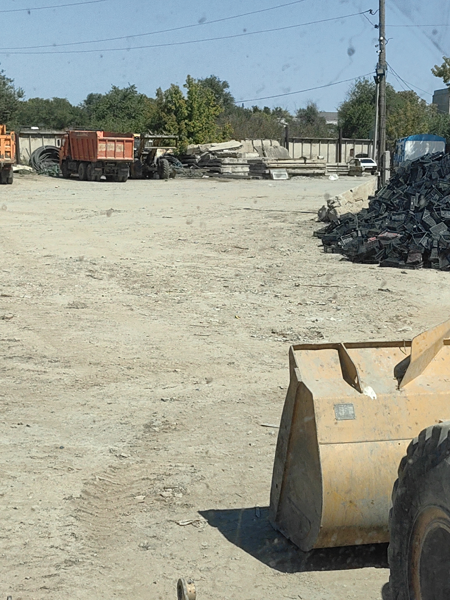 The image depicts an outdoor industrial or construction site. In the foreground, a yellow front-end loader with a large bucket is partially visible, along with a section of its tire. The ground is composed of dirt and gravel, with tire tracks evident. In the middle ground, an orange dump truck is parked, and behind it, a smaller construction vehicle, possibly a backhoe or skid steer, is also present. A pile of what appears to be industrial pipes or hoses is stacked to the left of the dump truck. Further back, there are stacks of concrete slabs and other building materials. A white car is parked near a building structure in the background, and a large pile of black plastic crates is situated to the right. A utility pole with electrical wires stands prominently in the background. The sky is clear and blue. The scene appears to be a typical day at a material storage or processing facility, possibly a construction yard or recycling center.
