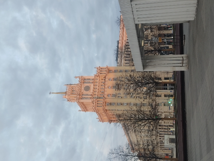 The image depicts the Ministry of Agriculture building, a Stalinist skyscraper in Moscow, Russia, taken from street level. The sky is overcast. A modern structure, possibly a bus stop or covered walkway, is to the right. The building's architecture features a spire, a clock, and decorative elements. Bare trees are visible in front of the building. A traffic light is visible in the background. The sidewalk is concrete.