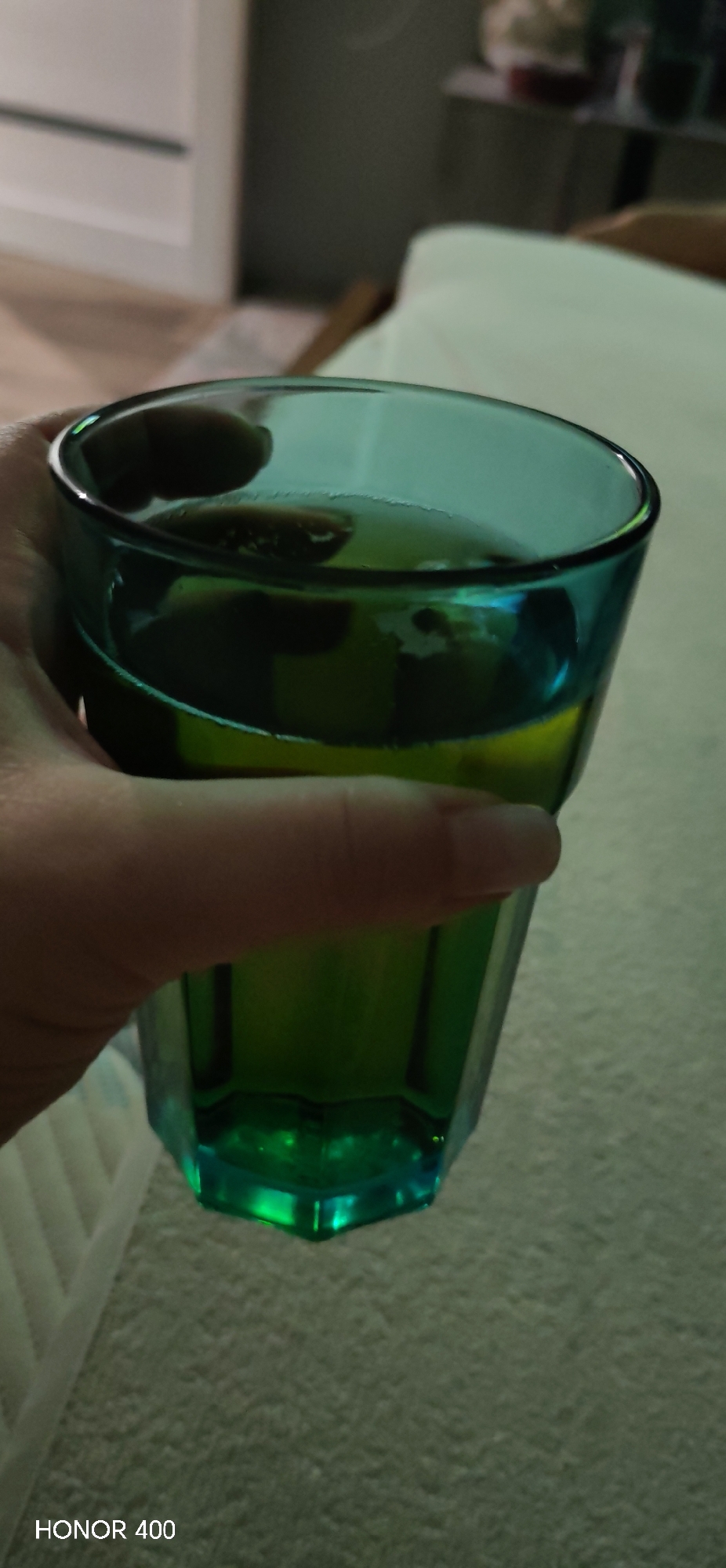 A close-up shot shows a hand holding a faceted, teal-colored glass filled with a yellowish-green liquid. Bubbles are visible on the surface of the liquid. The glass has a thick rim and a multifaceted base. A blurred background shows a light-colored surface, possibly a blanket or a couch. The bottom left corner of the image has the text "HONOR 400". No information suggests the image was taken in Noviy, Russia.