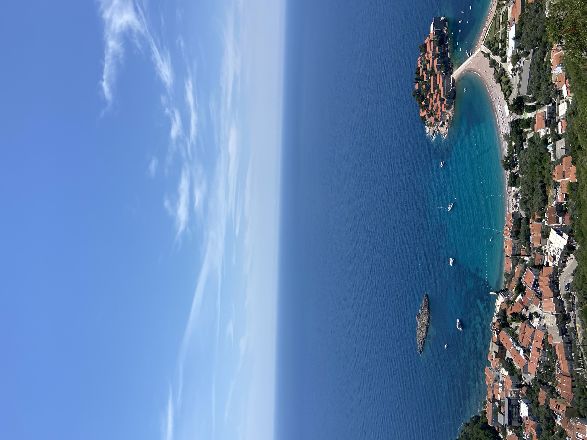 The image depicts a high-angle aerial view of Sveti Stefan, Montenegro, on a clear day. The scene is dominated by the vast expanse of the deep blue Adriatic Sea meeting a bright blue sky with scattered white clouds. In the foreground, a coastal town with red-tiled roofs stretches along the shoreline, nestled amongst green foliage. A prominent feature is the islet of Sveti Stefan, connected to the mainland by a narrow causeway. The islet is densely packed with buildings, also featuring red roofs, and surrounded by the turquoise waters of the bay. Several boats, including sailboats and smaller vessels, are anchored or sailing in the clear water. A sandy beach curves around the causeway, dotted with numerous sunbathers. The overall impression is of a picturesque Mediterranean coastal resort.