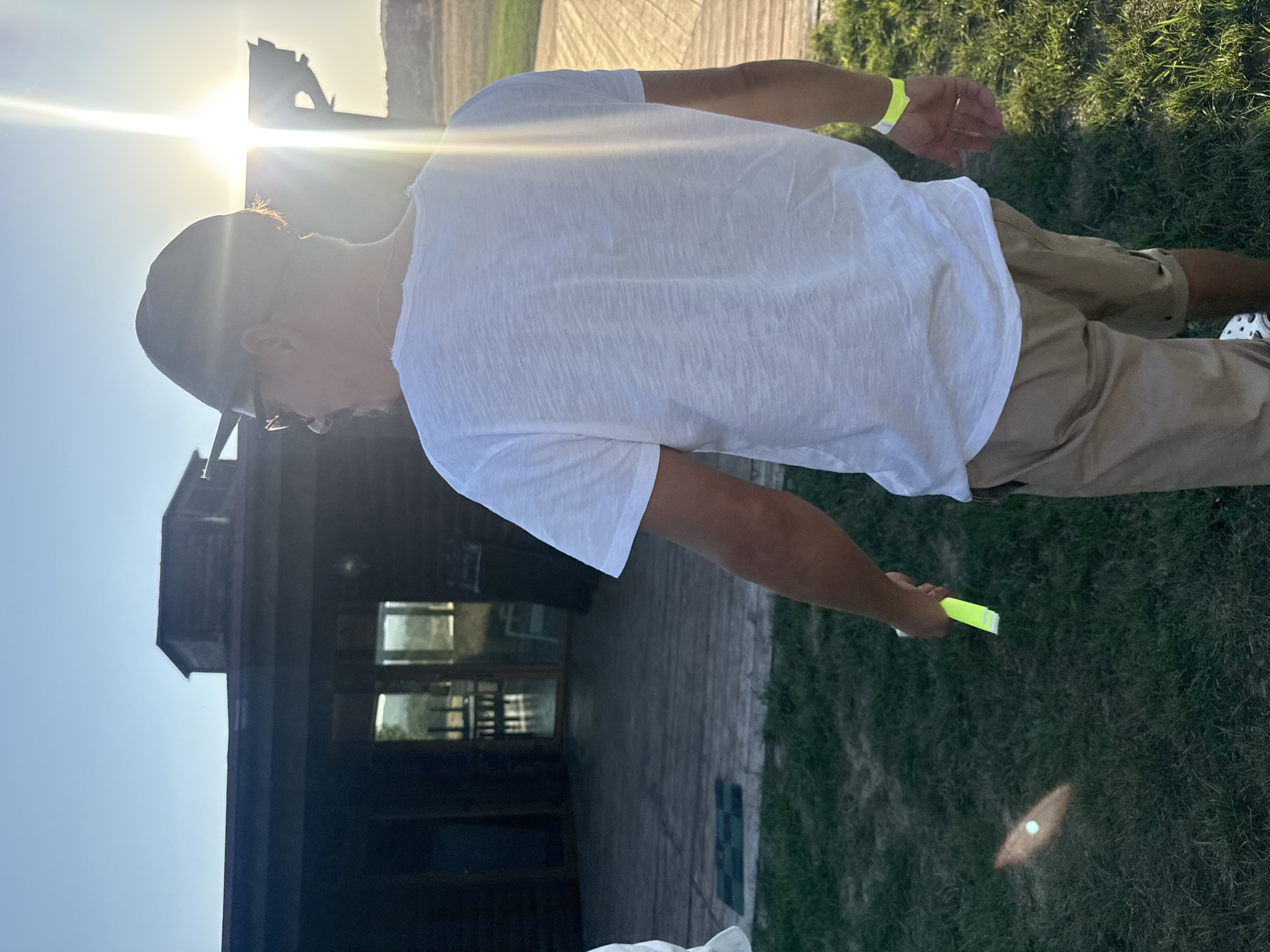 The image depicts a person from behind, wearing a white t-shirt, khaki shorts, a dark baseball cap, and sunglasses. They are holding a bright yellow object, possibly a marker or a light stick, in their right hand. The person is standing on a grassy area next to a wooden deck. In the background, there is a rustic wooden building with a porch. The sun is low in the sky, casting a strong glare and creating lens flare effects. The setting appears to be outdoors, possibly in a rural or recreational area, given the natural environment and the rustic architecture. The location is identified as Orlinovsky District, Ukraine.