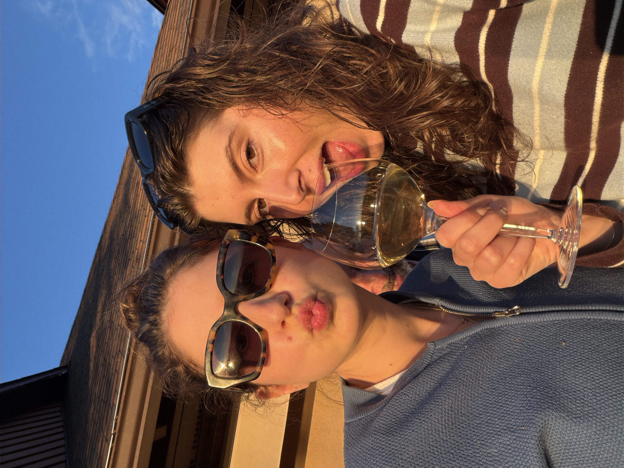 Two women are taking a selfie outdoors in front of a house with a brown shingled roof. The woman on the left is wearing a blue zip-up sweater and tortoise-shell sunglasses, with her hair in a bun. She is puckering her lips. The woman on the right is wearing a striped sweater and black sunglasses perched on her head. She is drinking from a wine glass, with her tongue extended. There is a man visible in the background between the two women. The sky is clear and blue, and the setting sun casts a warm glow on the scene. The location is Mont-Tremblant, Canada.