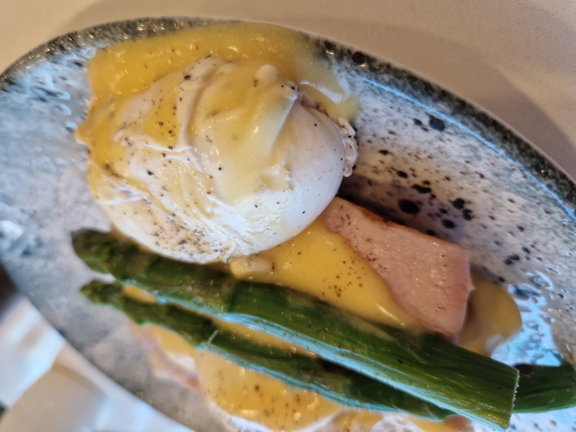 A close-up shot shows a plate of Eggs Benedict, a breakfast dish. The dish consists of two poached eggs on top of ham and asparagus, all covered in hollandaise sauce. The plate is grey and speckled with black. The background is blurred but suggests an indoor setting. The image does not provide any information about the city of Sofia, Bulgaria, or any people.