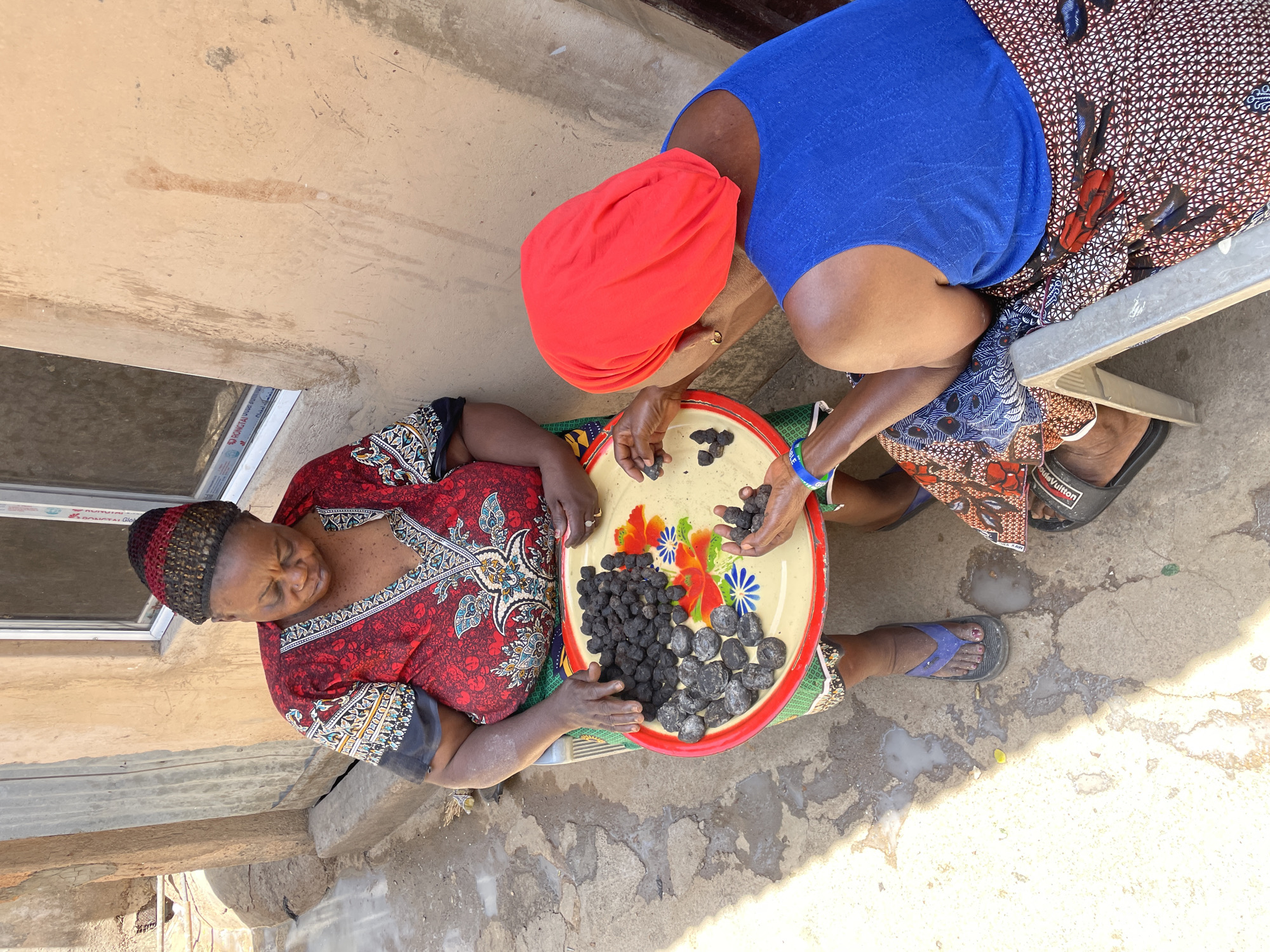 This high-angle, outdoor photograph, taken in Jos, Nigeria, captures two women engaged in an activity with a tray of dark, solid items under bright daylight, indicating a sunny day.
On the left, a woman is seen from the chest up, wearing a red and black patterned tunic with intricate designs and a dark knitted hat. She is looking downwards, presumably at the tray. To her right, a second woman, dressed in a royal blue top, a red headwrap, and a patterned wrapper skirt, is seated on a low, light-colored stool. She is actively handling the dark items, holding a small pile in her hands, suggesting sorting or preparation.
A large, circular yellow tray with a red rim dominates the center foreground, beautifully decorated with colorful floral patterns in red, orange, blue, and green. The tray holds numerous dark, irregularly shaped objects; some are rough and black, while others appear smoother, rounder, and greyish. These items could be charcoal briquettes or a type of processed food product.
The scene is set on a concrete ground, showing some stains and marks of wear. In the background, part of a building wall and a window frame are visible. Text on the window frame includes "DONGTAL GLOB..." and "ALUMINIUM". The woman on the right wears black slippers, one of which has "Louis Vuitton" printed on it. The overall impression is one of focused work, possibly in a domestic or informal market setting.