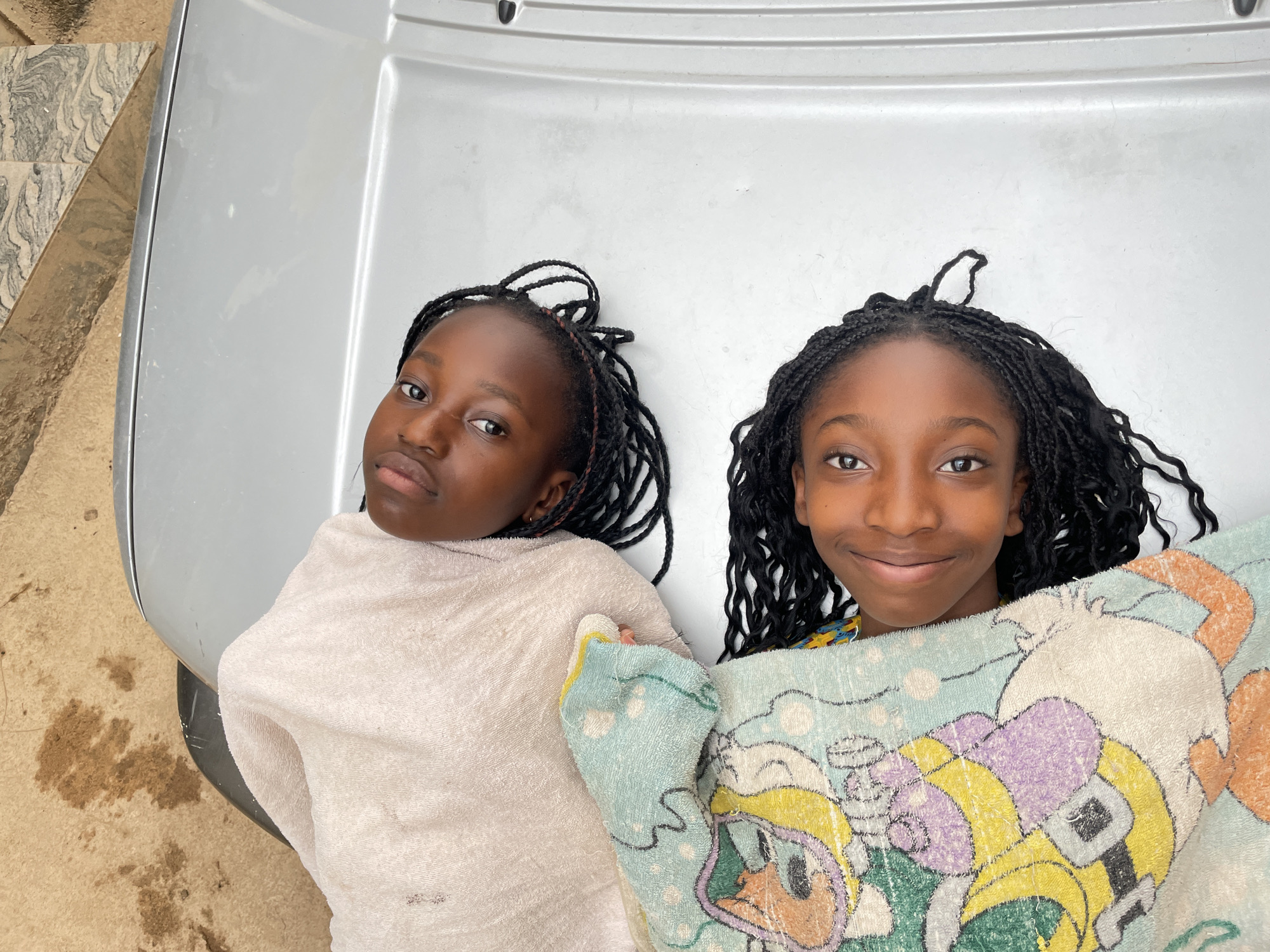 Two young, dark-skinned girls lie on the hood of a silver car. The girl on the left is wrapped in a white towel, while the girl on the right is covered with a blue and yellow cartoon-patterned towel. Both girls have their hair in braids. The location appears to be Jos, Nigeria, based on the physical features of the individuals and the environment. The background includes a textured stone surface and a concrete ground surface.