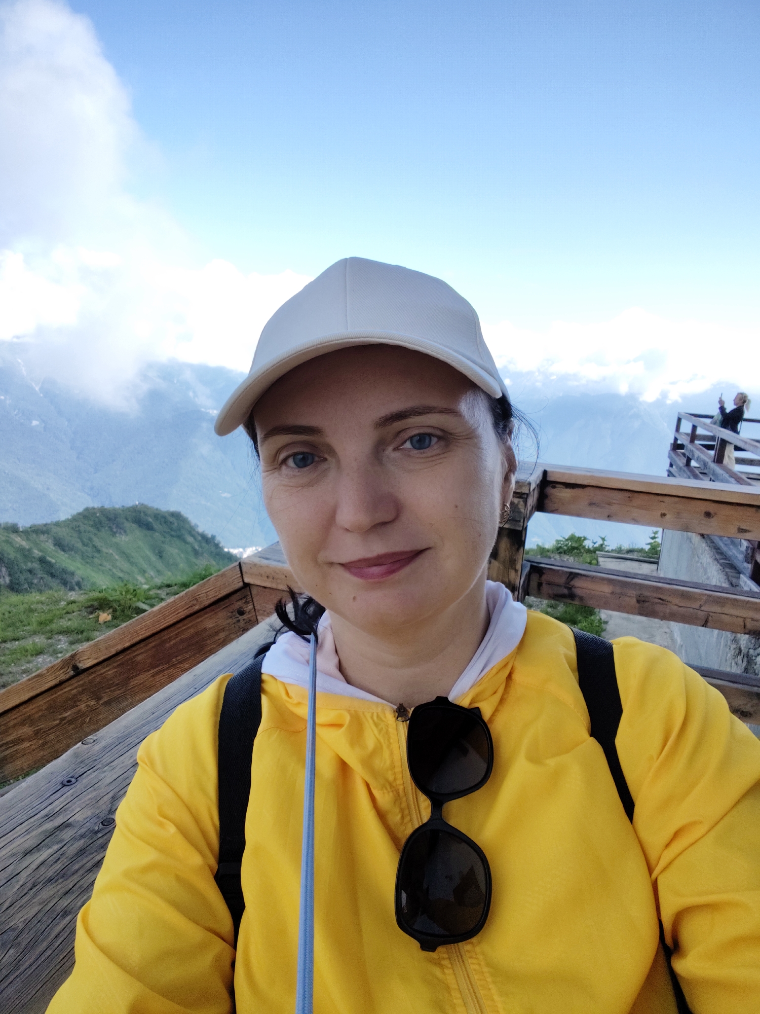 A selfie of a woman wearing a white baseball cap and a bright yellow jacket with a white hood is taken on a wooden observation deck. She has blue eyes and fair skin with some freckles. Black sunglasses are hooked onto the front of her jacket. In the background, a person can be seen on a wooden staircase. The setting is a mountainous landscape with lush green slopes and a bright blue sky with scattered clouds, consistent with a location in Estosadok, Russia.