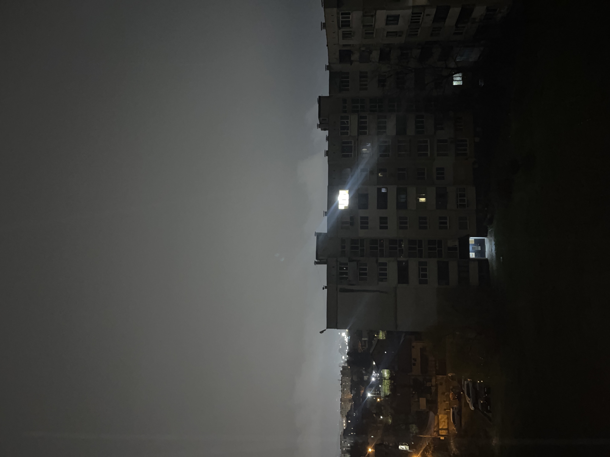 A nighttime panoramic view of a residential area in Sofia, Bulgaria, characterized by low visibility due to an overcast sky. The upper third of the image is dominated by a dark, gray sky with visible plumes of smoke or exhaust rising from the urban landscape below. The midground features a prominent, multi-story concrete residential building, approximately 8-10 stories tall, with numerous windows. Several windows on this building are illuminated, most notably a single, intensely bright window on an upper floor of the left-center section, casting a distinct light flare. The building's facade shows evidence of age and dark stains. At the ground level of this building, a brightly illuminated entrance with a blue door is visible. To the left of the main building, a street or parking area is partially visible under orange streetlights, illuminating at least three parked vehicles, including a light-colored SUV and darker sedans. Distant city lights create a glowing horizon beneath the low cloud cover in the far background. No individuals are discernible within the frame.