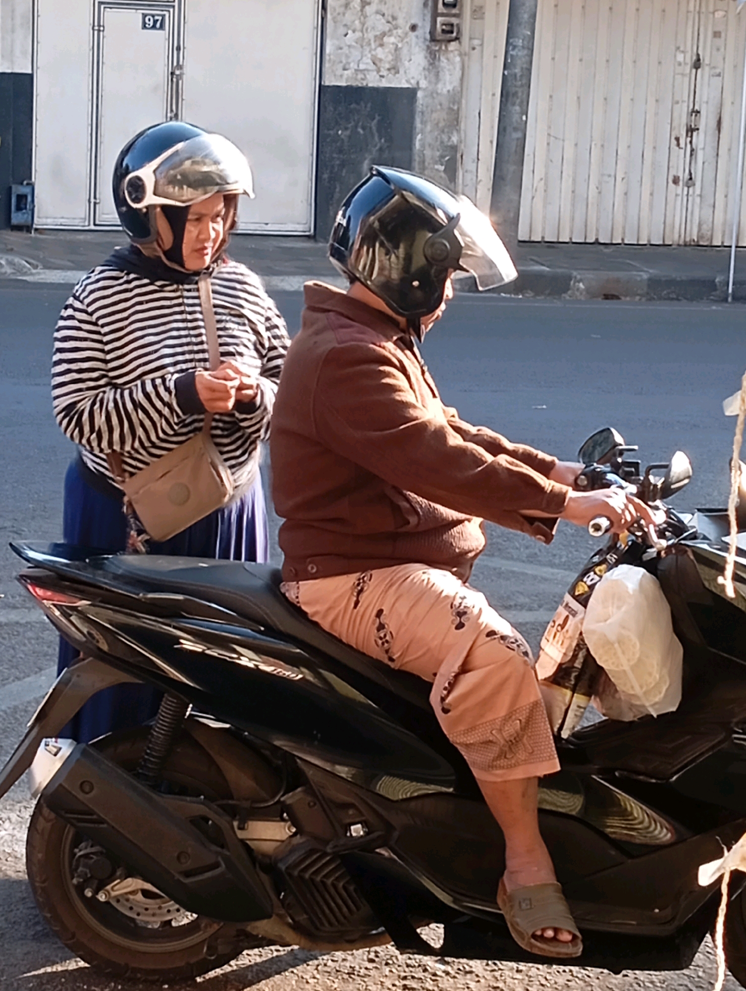 Two people, likely women, are seen on a black scooter parked on a paved street. The person in the foreground is wearing a brown jacket and light-colored pants with a subtle pattern, and is seated on the scooter, holding the handlebars. The person behind them is standing and wearing a striped long-sleeved shirt and a blue skirt, with a crossbody bag. Both are wearing helmets. The background shows a building with white doors and the number "97" visible above one door, suggesting an urban environment in Malang, Indonesia. The lighting suggests it is daytime.