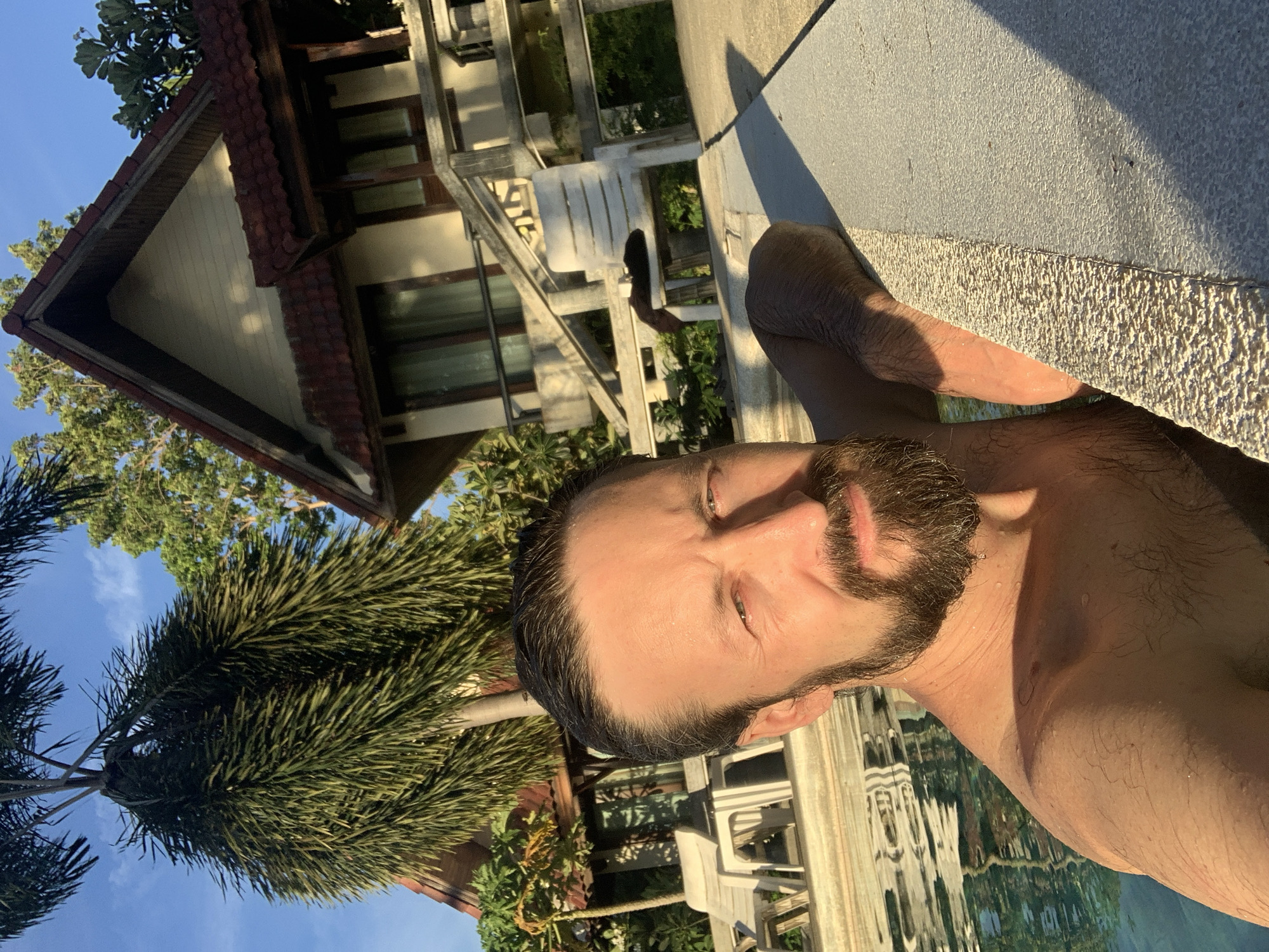A man with dark hair and a beard is in a swimming pool in front of a tropical resort. He is leaning on the edge of the pool, with the water reflecting the sunlight. Behind him, two bungalows with red tile roofs and wooden balconies are visible, surrounded by lush green trees and palm trees. The sky is clear blue with a few scattered clouds. The scene appears to be set in Baan Bang Rak, Thailand, likely during the daytime.