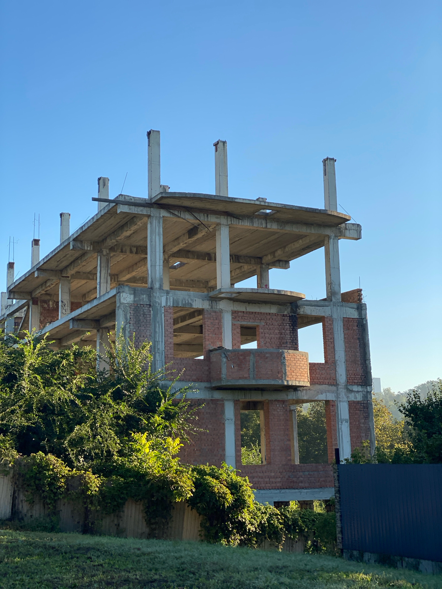 An unfinished, multi-story concrete and brick building stands under a clear blue sky, partially obscured by lush green foliage. The building features exposed concrete floors and pillars, with brick walls forming the lower levels and sections of upper floors. Windows are cut out of the brickwork, revealing the empty interior structure. Dense green bushes and vines grow along a low fence in the foreground, with a dark metal fence visible on the right side of the image. The overall scene suggests a construction site in a suburban or semi-rural area.