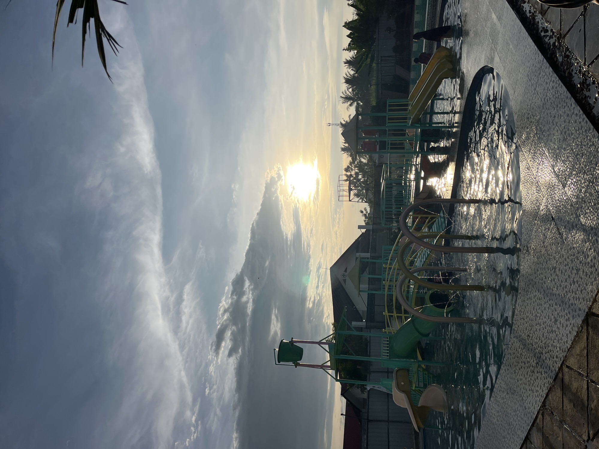 The image depicts a children's water park in Lhokseumawe, Indonesia. The photo is taken at ground level near the edge of a pool with tile trim. The water park has a small slide, a water dumping bucket, and a climbing frame structure, all situated in a pool of water. Sunlight reflects brightly off the surface of the water, creating glare.
Two people are visible in the background of the image, standing near another water slide. Trees and buildings are also visible in the distance, silhouetted by the bright sun. The sky is filled with clouds.