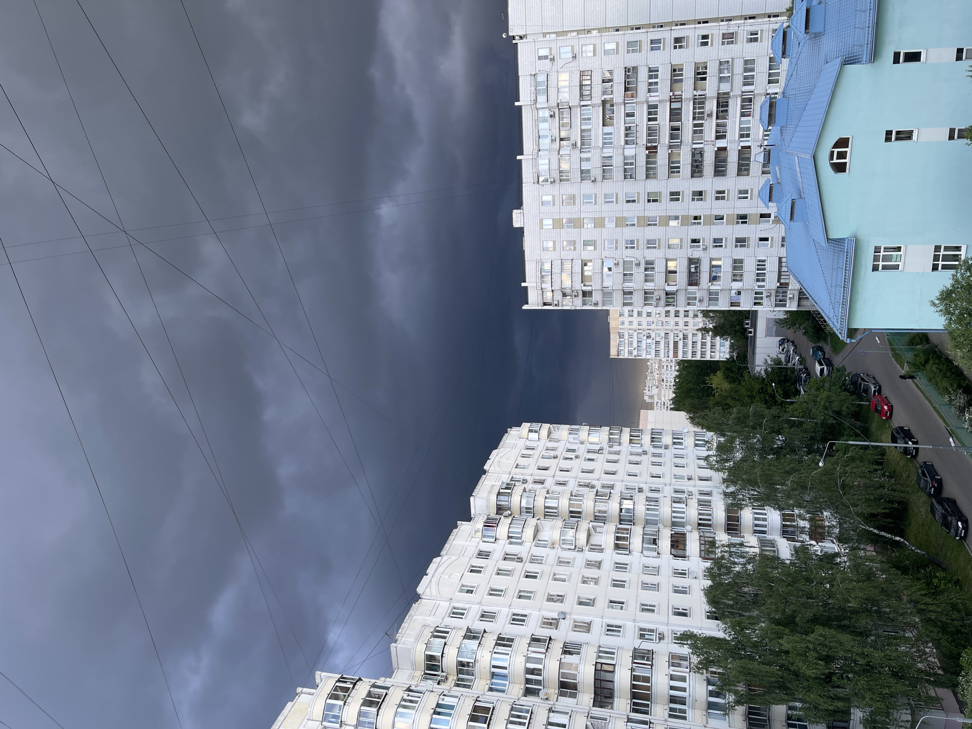The image depicts a cityscape in Moscow, Russia, showcasing a dramatic sky with dark, ominous storm clouds looming between tall, white apartment buildings. Overhead power lines crisscross the sky. To the right, a building with a distinctive blue roof is visible. Cars are parked along a street lined with trees below. The overall atmosphere suggests an impending storm in an urban setting.