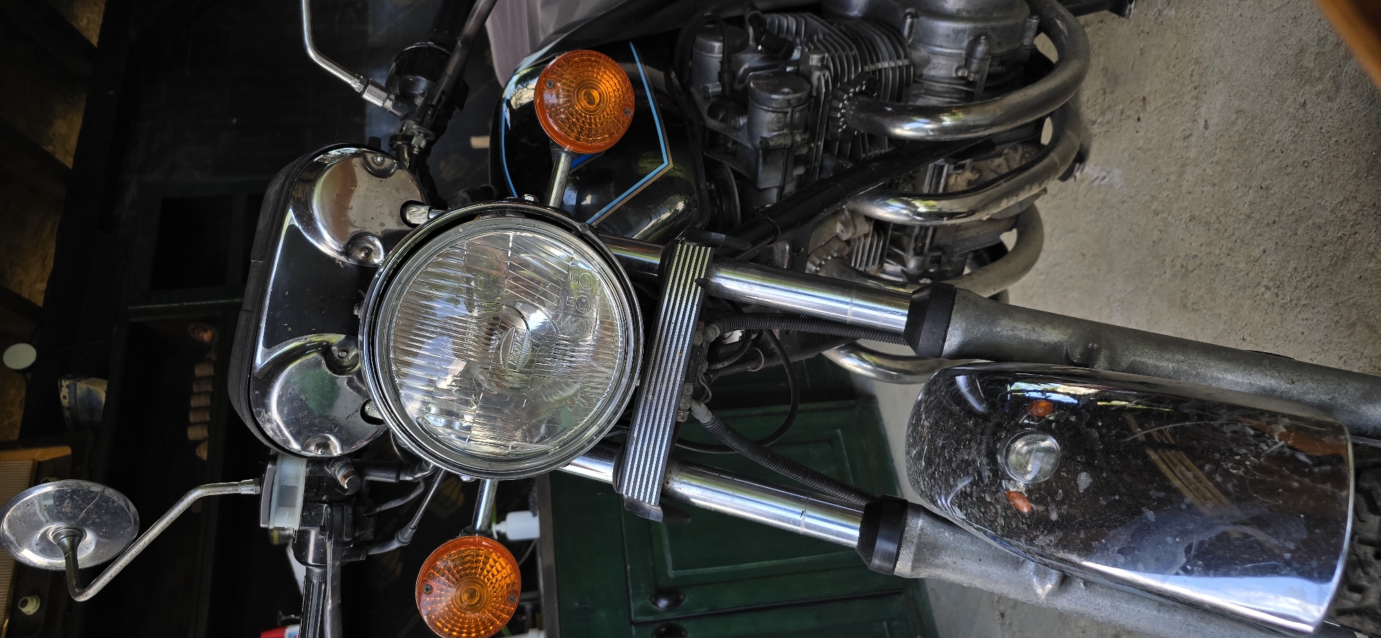 The image is a close-up, eye-level, vertical shot of the front of a vintage motorcycle parked outdoors. The motorcycle is predominantly black and chrome. The focal point is the large, circular headlight with a clear lens and a chrome bezel, positioned centrally. To the left and right of the headlight are orange turn signals. The motorcycle's handlebars and mirrors are visible at the top of the frame, along with some of the engine components and exhaust pipes. The front fender is dark and appears to be somewhat dirty or smudged. The background consists of dark green cabinetry or walls and a dark, covered area above. The ground beneath the motorcycle is a light-colored concrete surface. There are no people present in the image. The scene appears to be in an outdoor setting, possibly a garage or a sheltered area.
