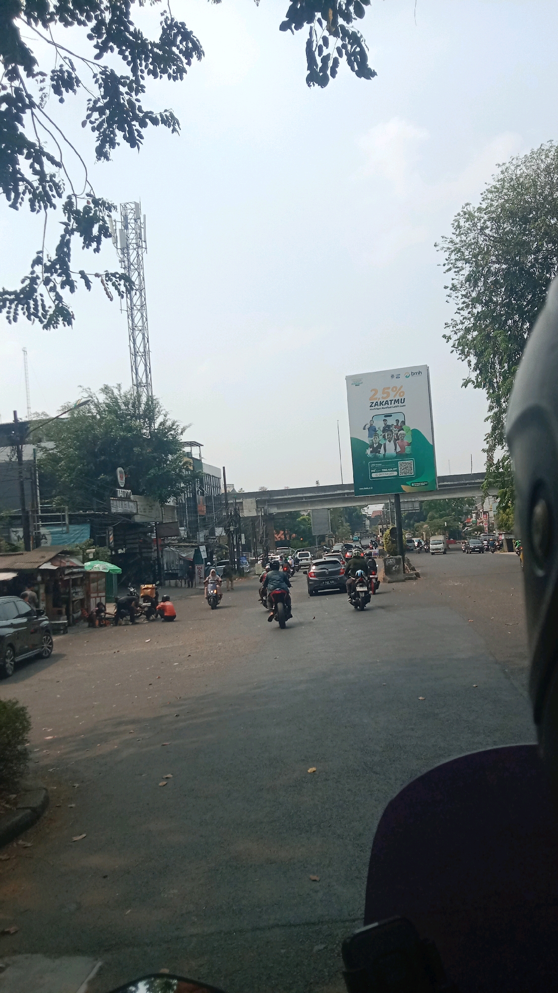 The image depicts a street scene in Bekasi, Indonesia, on a hazy day. A busy road is visible with several motorcycles and cars navigating through traffic. On the right side of the road, a large billboard advertises "25% ZAKATMU" with an image of people and a mobile phone app, suggesting a religious or charitable campaign. In the background, an elevated highway structure and a telecommunications tower can be seen, indicating urban infrastructure. On the left side of the road, there are small shops and stalls, possibly selling food or goods, with some people sitting around. The perspective suggests the photo is taken from a moving vehicle, possibly a motorcycle, as a helmet is partially visible in the foreground. The environment appears to be a commercial or mixed-use area with moderate to heavy traffic.