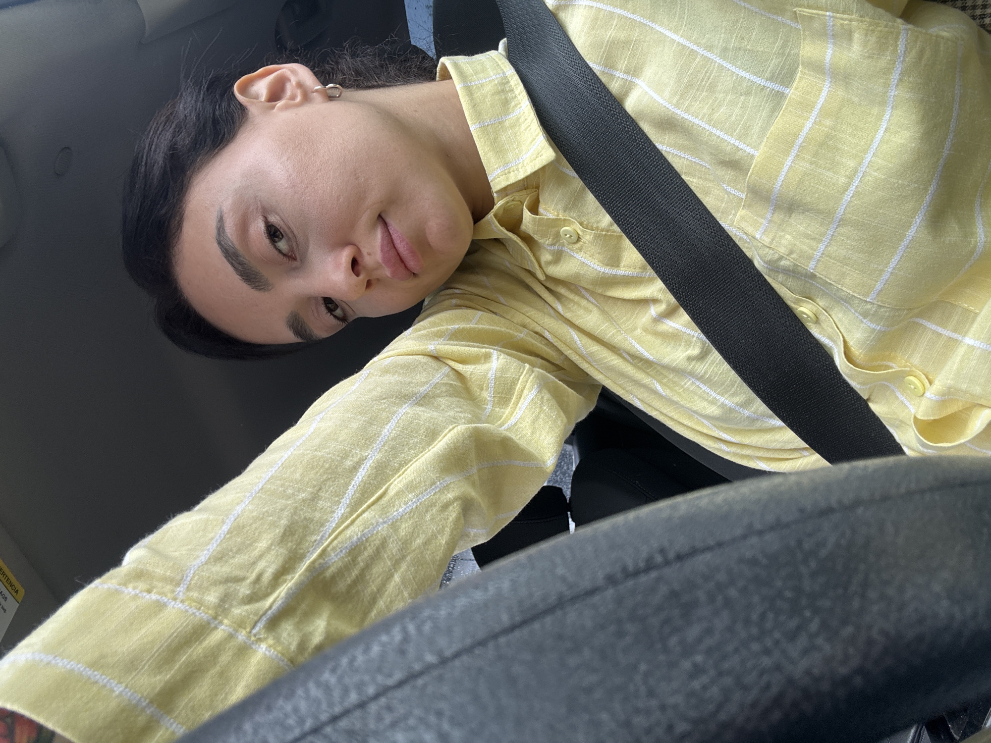 A person is seated in a vehicle, wearing a yellow and white striped button-down shirt and a seatbelt. Their face is visible, and they appear to be in a car. The steering wheel is in the foreground, partially obscuring the view. The location is likely Tbilisi, Georgia, inferred from the content, though not explicitly visible in the image.