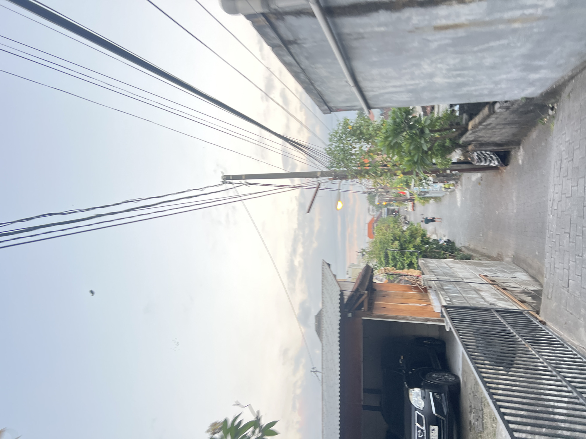 The image depicts a narrow street in Denpasar, Indonesia, at dusk. The sky is a pale blue with hints of pink and gray clouds. Overhead, numerous electrical wires crisscross the frame, supported by a dark utility pole. On the left, a black SUV is parked under a covered carport, next to a building with a corrugated metal roof and wooden walls. A metal fence separates the carport from the paved street. The street itself is lined with gray concrete walls on the right and a mixture of walls and vegetation on the left. Further down the street, a single person is walking away from the camera. The street is paved with cobblestones or similar material. The overall impression is of a residential area in a densely populated urban environment.