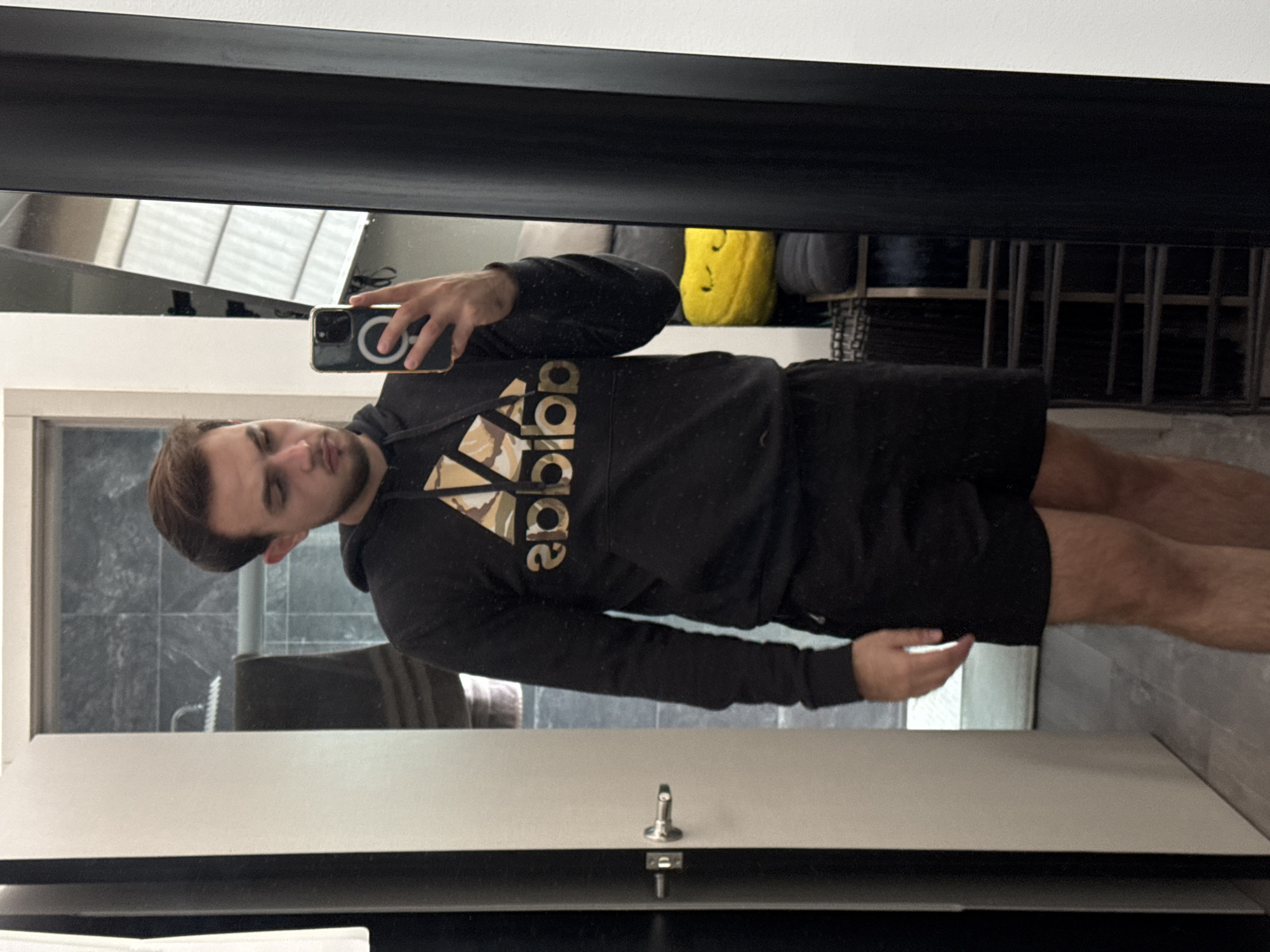 A man is taking a selfie in a mirror. He is wearing a black Adidas hoodie and black shorts. The reflection shows a glimpse of a room with a window and some items on a shelf. The location in the city of San Bernardino Tlaxcalancingo, Mexico is not discernible from the image.