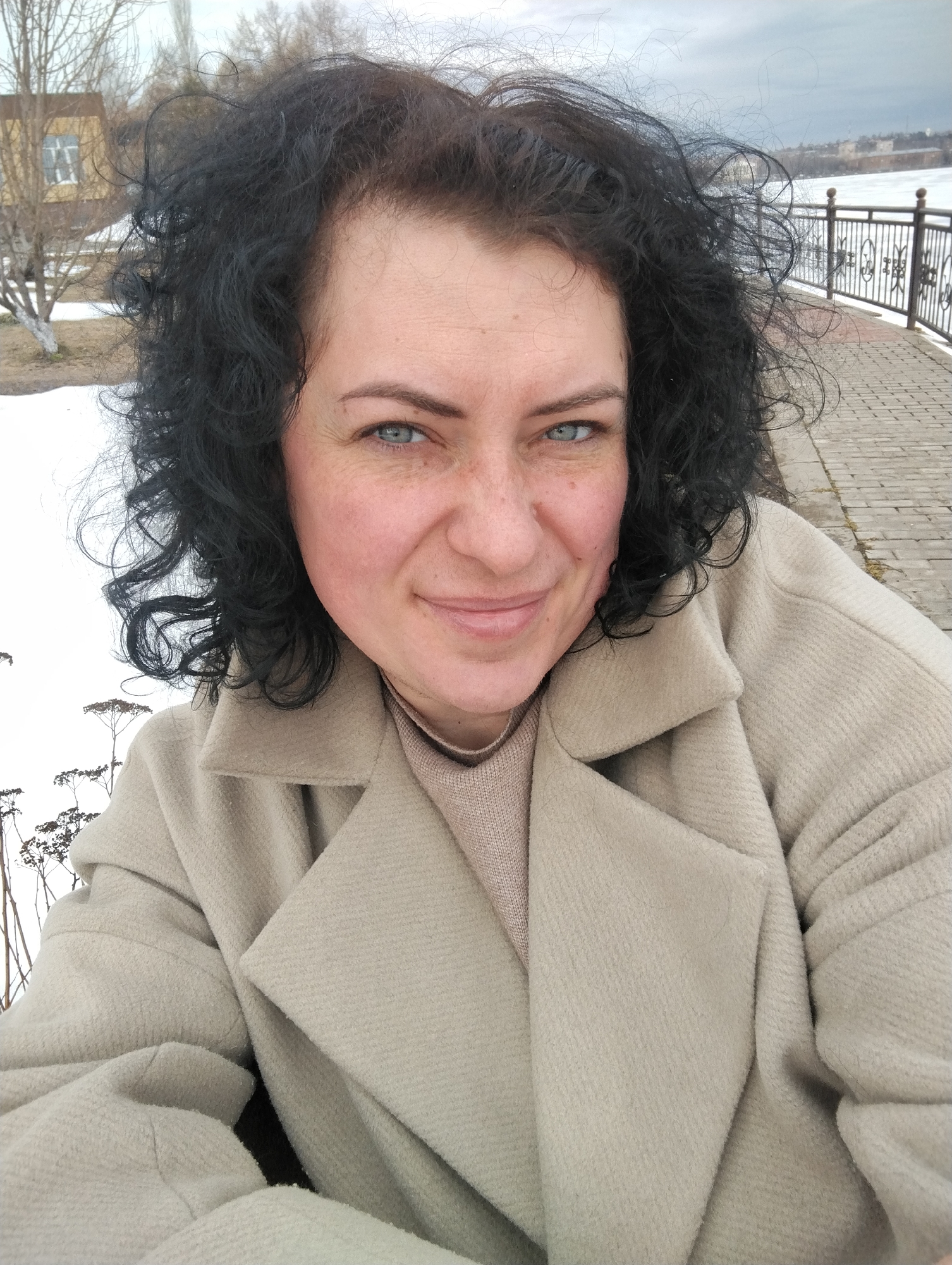 A medium close-up selfie shows a woman outdoors on a bridge with snow visible on the ground. She has fair skin, blue eyes, and dark, curly hair. She is wearing a beige coat and a light brown turtleneck sweater. The bridge is made of brick and has a black metal railing on the right side. In the background, there is a partially visible building, possibly a house, and some trees. A body of water is also visible behind the railing. Considering the presence of snow, the style of the architecture and landscape elements, it is likely that the location could be a bridge in Votkinsk, Russia, although exact verification cannot be definitively made without further information to pinpoint location.