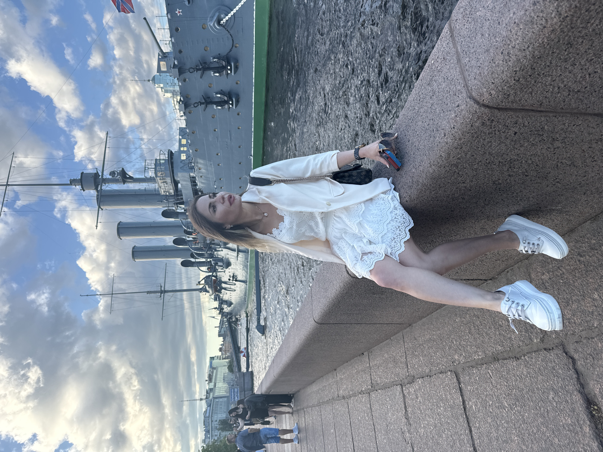 A woman is sitting on a stone embankment with a large grey warship behind her in Saint Petersburg, Russia. The woman has long dark hair and is wearing a white lace dress and a cream-colored blazer. She is looking to her left. In her right hand, she holds sunglasses and what appears to be a mobile phone. The warship, the Aurora cruiser, is docked on the Neva River. The sky is cloudy.