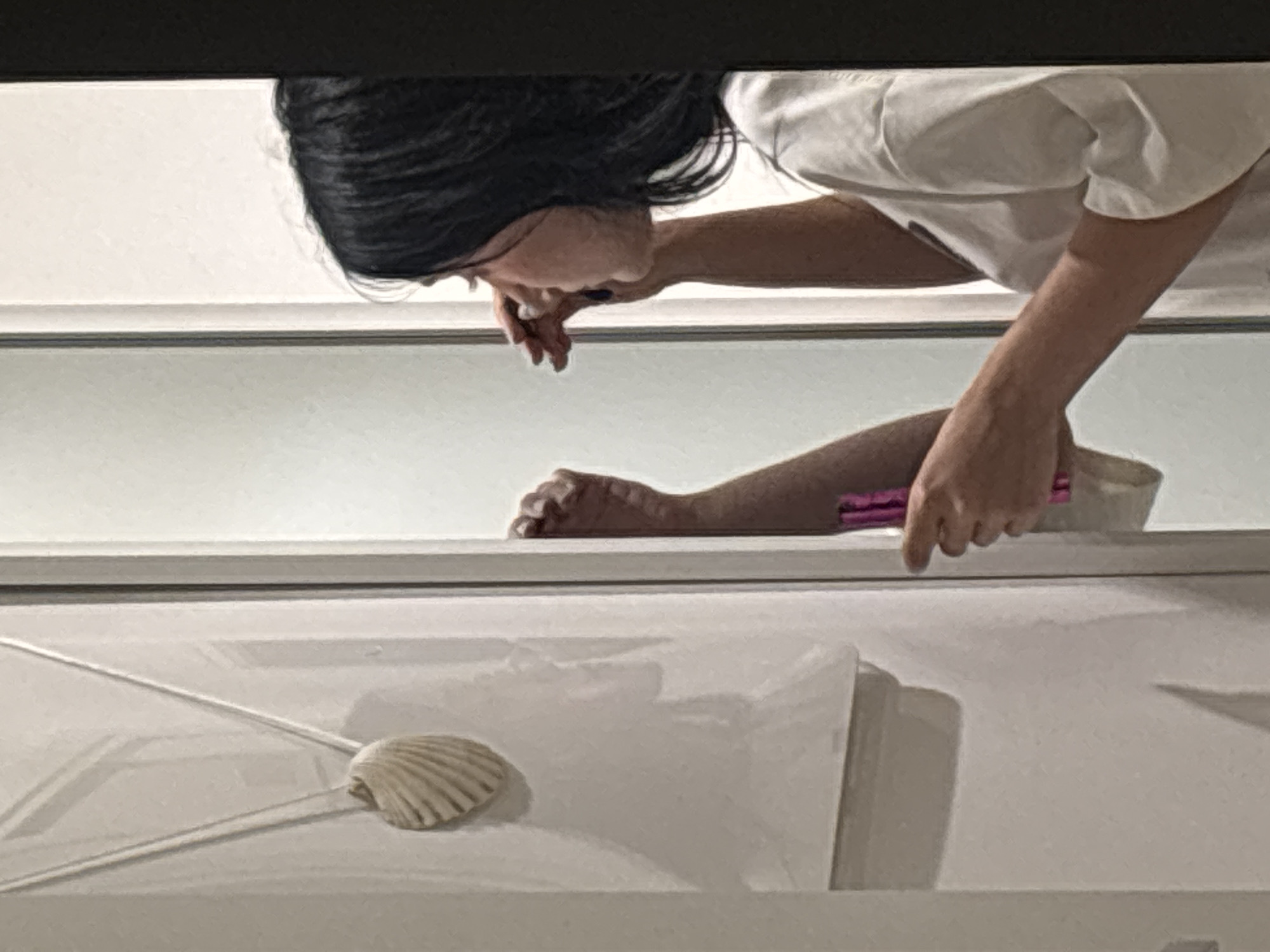 A medium shot captures a young woman with dark hair applying makeup in front of a mirror. She is wearing a white t-shirt. Her left hand holds a pink cosmetic applicator, and her right hand is near her face. The mirror reflects a shell necklace hanging on a wall in the background. The image is taken indoors, likely in a residential setting, as evidenced by the decorative shell and the simple interior design. The overall impression is that of a candid moment of personal grooming. This scene is not located in the city of Cupra Marittima, Italy.
