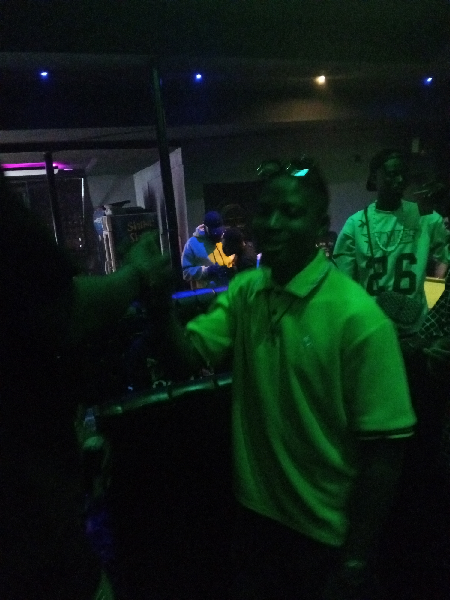 Here is a concise, forensic description of the scene:
**Location:** Unknown, Unknown. Indoor environment, possibly a nightclub or performance venue.
**People:** Multiple individuals are visible. One person is in the foreground, wearing a green polo shirt and sunglasses on their head. Another person is behind them, wearing a green long-sleeved shirt with the number "26" on it and a hat. Further in the background, another person is wearing a blue hoodie.
**Objects:** A DJ setup is visible behind the foreground individual. "SHINE" is printed on the DJ stand.
**Actions and Interactions:** A handshake or high-five appears to be taking place between the foreground individual and an unseen person partially blocking the view on the left. People in the background appear to be enjoying the event.
**Environment:** The scene is dimly lit with blue and green ambient lighting.