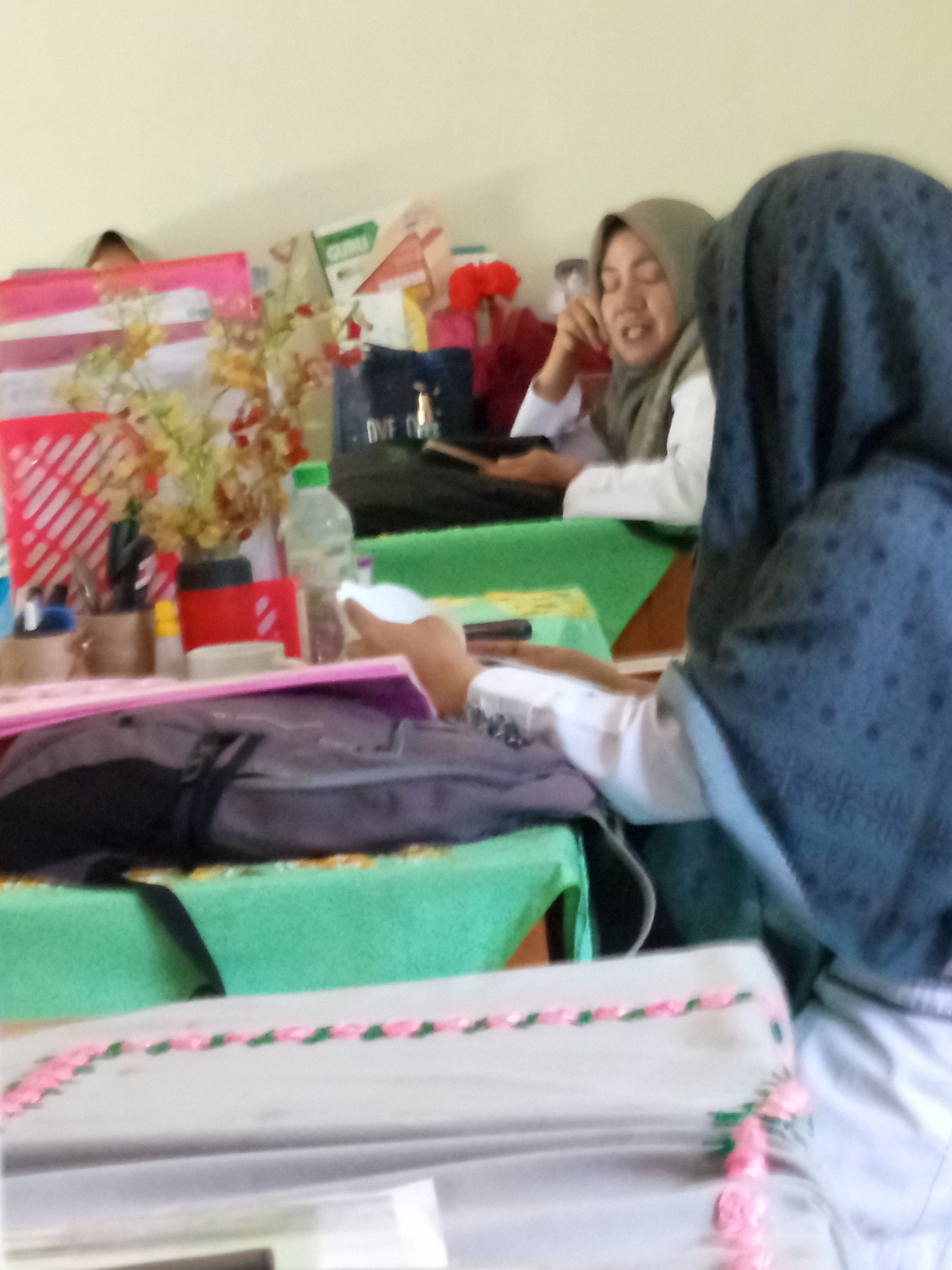 In Lumutan, Indonesia, two individuals are seated at a table. The individual in the foreground, facing away from the camera, is wearing a dark hijab with a dotted pattern. The person behind them, facing the camera and wearing a light green hijab, is smiling and looking down at something on the table. The table is covered with a green cloth and displays various items including a pink folder, a red container, pens in a holder, a bottle of water, and a dark colored backpack. The background shows a wall and some papers with writing on them. The scene appears to be indoors, possibly in a classroom or office setting.