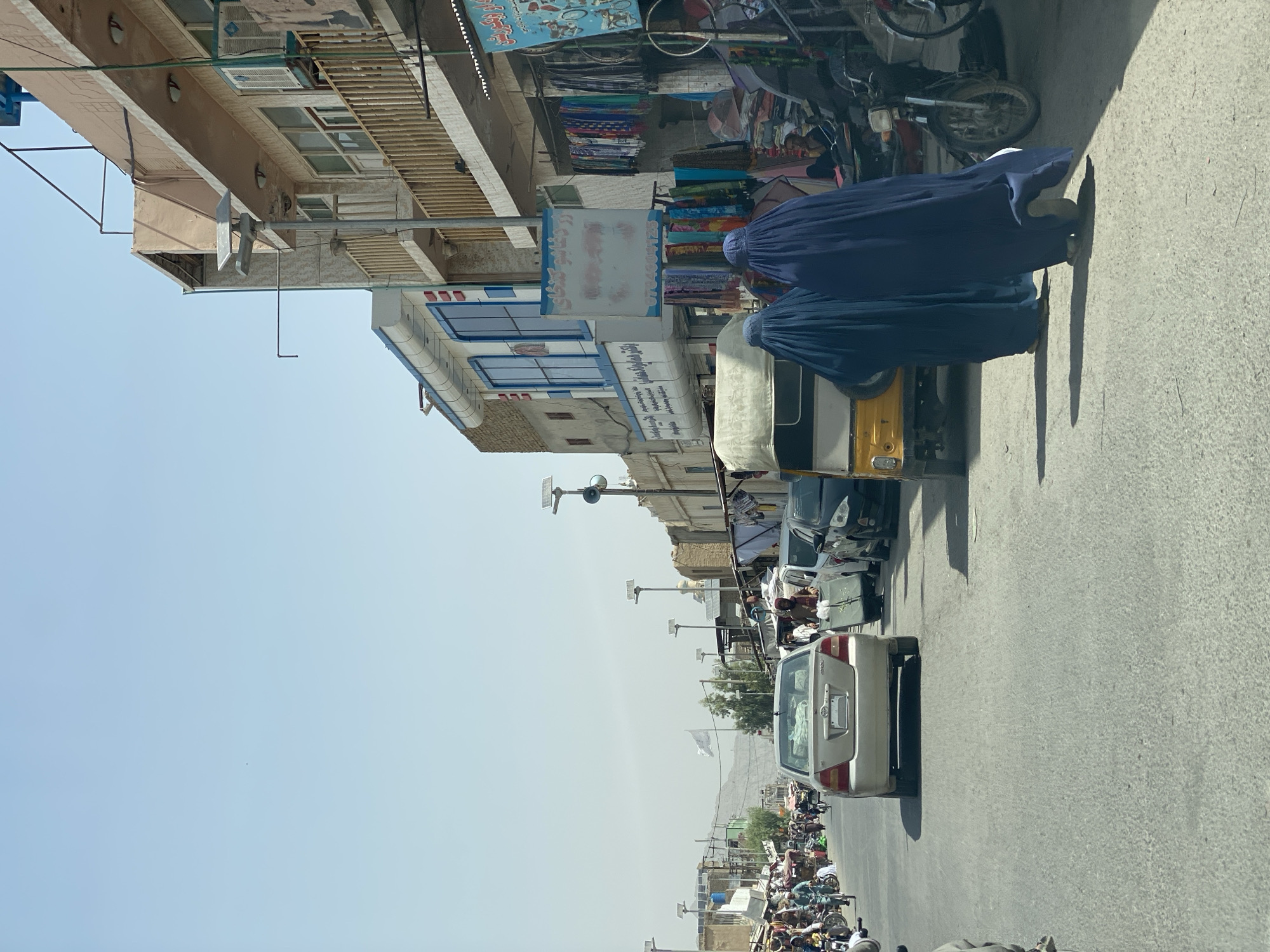 A daytime, eye-level, outdoor street scene in Kandahar, Afghanistan. The image is captured from a moving vehicle and shows a bustling street lined with shops and commercial buildings. In the foreground, two women are walking away from the camera, both wearing dark blue burqas. Behind them, a yellow auto-rickshaw is visible. The street is paved with asphalt and shows signs of wear.
Vehicles are present on the street, including a silver sedan in the middle ground, and several other cars and motorcycles on the right side of the road. Shops on both sides of the street display goods, including textiles and clothing, hanging outside. One building on the right features a balcony with wooden railings and an air conditioning unit. On the left side of the street, more shops and people are visible, with some individuals on motorcycles.
The sky is clear and bright blue. Solar-powered streetlights are mounted on poles along the street, indicating modern infrastructure integrated into the urban environment. A white flag with an emblem is visible on a flagpole in the distance, positioned against a backdrop of a dusty, mountainous landscape. The overall impression is a vibrant, active marketplace in a busy urban setting.