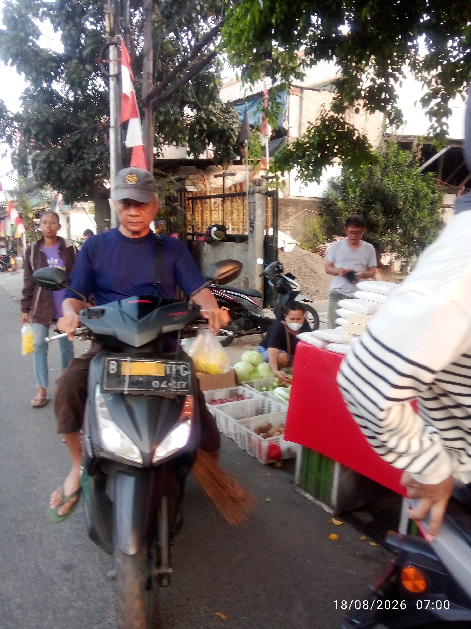 A man wearing a blue t-shirt and a gray baseball cap rides a black motorcycle on a street in East Jakarta, Indonesia. He has a dark bag slung across his chest. To his left, a woman wearing a purple t-shirt and a brown jacket walks along the sidewalk, carrying a yellow plastic bag. In the background, a man in a white t-shirt stands behind a stall selling produce, which includes white bags of rice, green cabbages, and red onions. A motorcycle is parked behind him, and a pile of gravel is visible further back. The street is paved, and trees line the area. A red and white Indonesian flag is visible on a pole to the left of the frame. The timestamp on the image indicates "18/08/2026 07:00".