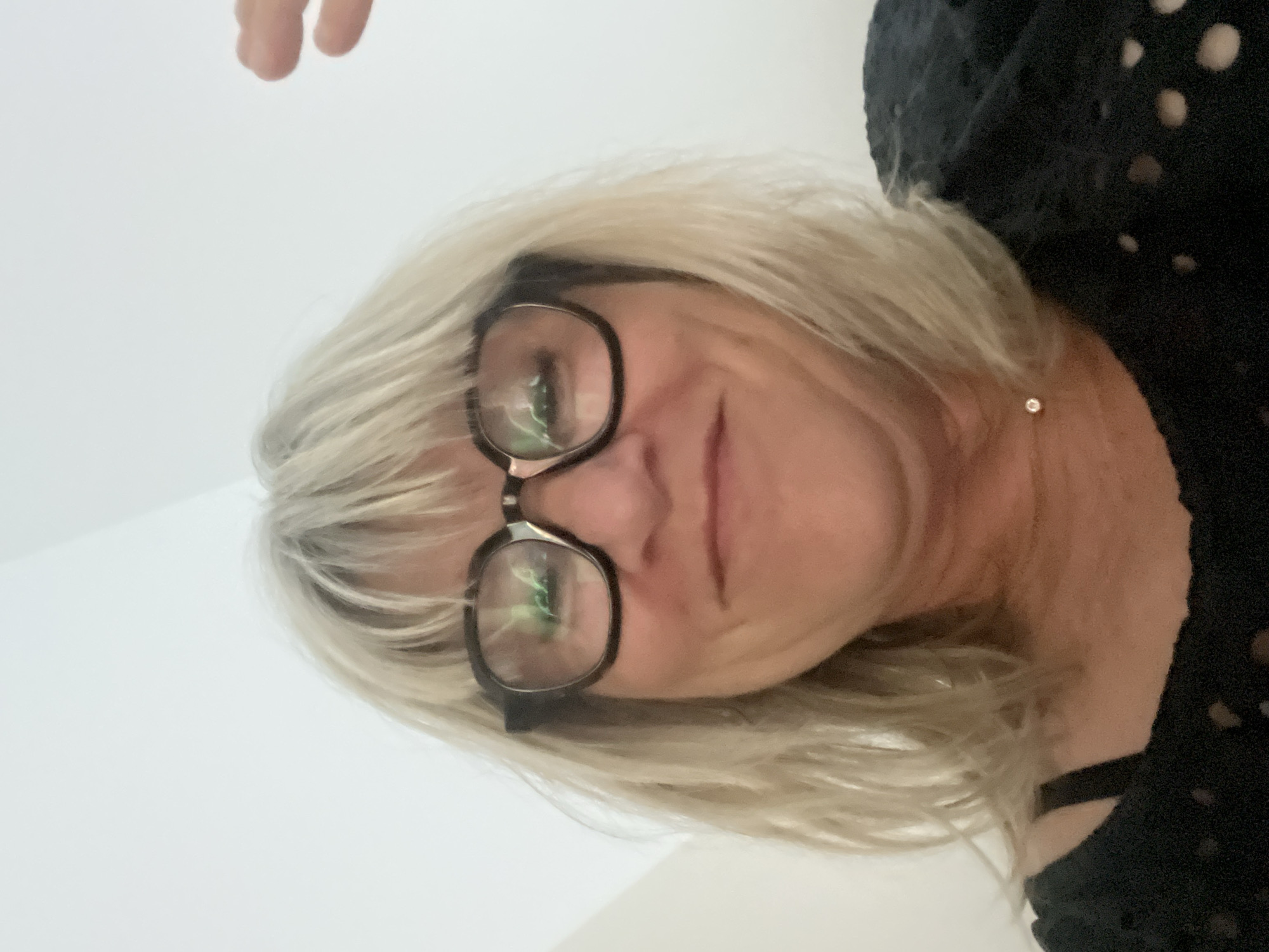 The image is a close-up, slightly upward-facing selfie of a light-skinned woman with blonde, shoulder-length hair and bangs. She is wearing dark-rimmed glasses and a black, patterned top with a bra strap visible. A small, light-colored pendant is visible on her neck. Her right hand is partially visible in the upper right corner of the frame, raised as if waving. The background is a plain, off-white wall or ceiling. There is no indication of the location being in Chambœuf, France.