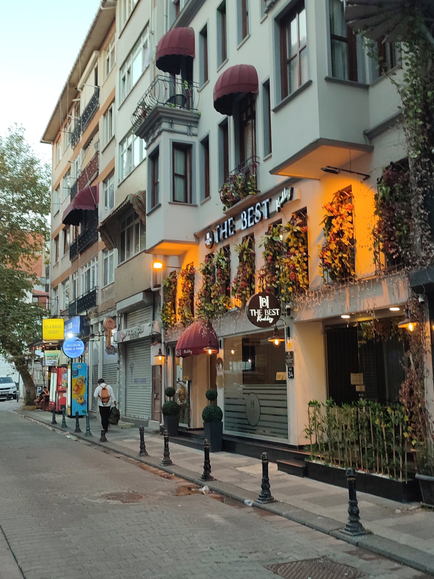 A low-angle, eye-level, outdoor, full-shot photograph captures a street scene in Kadıköy, Turkey. The buildings lining the street are multi-story structures with various businesses on the ground floor. The building on the right features a facade with arched awnings and decorative plants. The sign above its entrance reads "THE BEST" in large illuminated letters. Another circular sign below it also says "THE BEST" with "Kadikoy" underneath. The street is paved with cobblestones and has a raised sidewalk. Several black bollards line the edge of the sidewalk. A person wearing a white shirt, dark pants, and a backpack is walking away from the camera down the street. The sky is overcast, and there are trees on the left side of the image. Several signs in Turkish are visible on the buildings. The overall atmosphere suggests a commercial street in an urban environment.