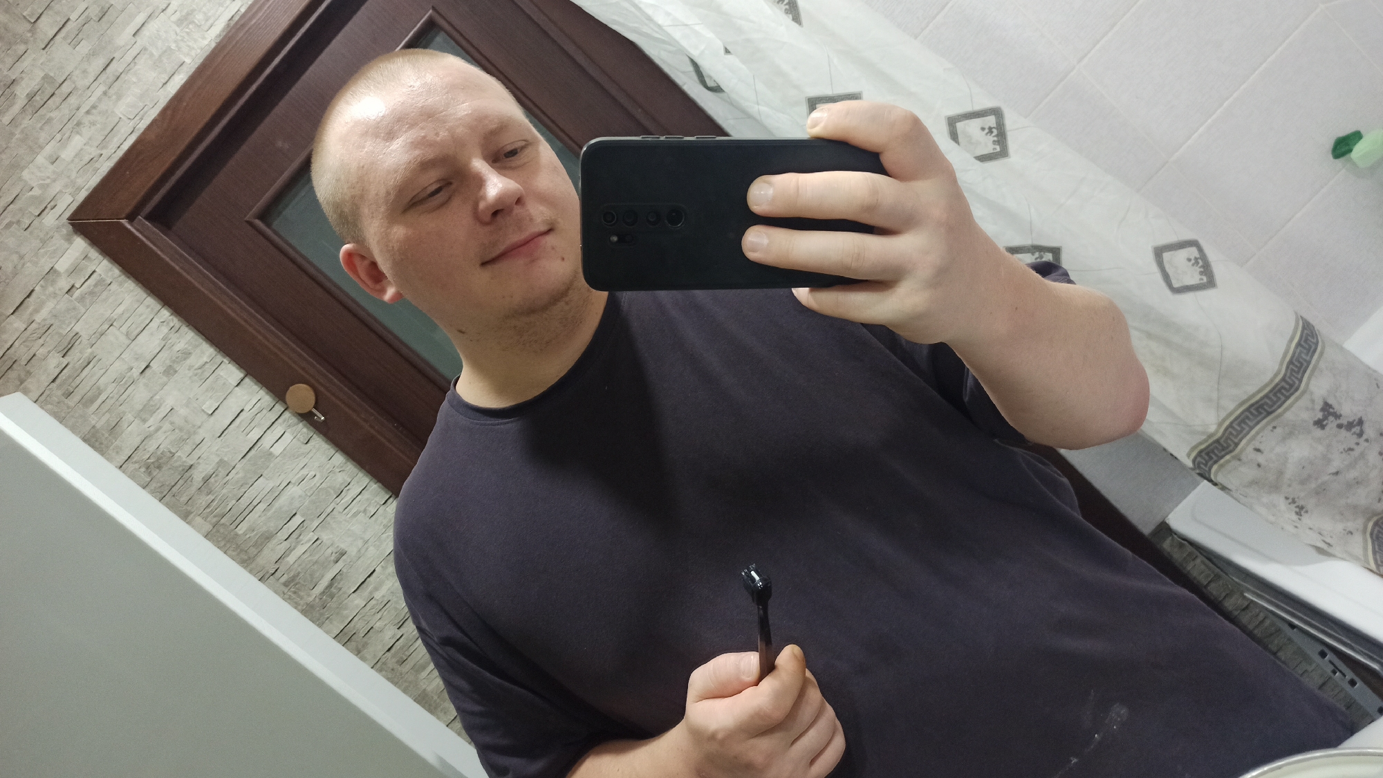 A bald Caucasian man with fair skin and light stubble is taking a selfie in a bathroom. He is wearing a dark gray t-shirt and is holding a black smartphone in his right hand, which is angled to capture his reflection. In his left hand, he holds a dark brown object with a small black roller on the end, possibly a microneedle derma roller. The background of the bathroom features a textured gray wall with vertical stone-like patterns and a dark brown wooden doorframe. A white shower curtain with a Greek key pattern is visible to the right. The lighting suggests it is indoors.
