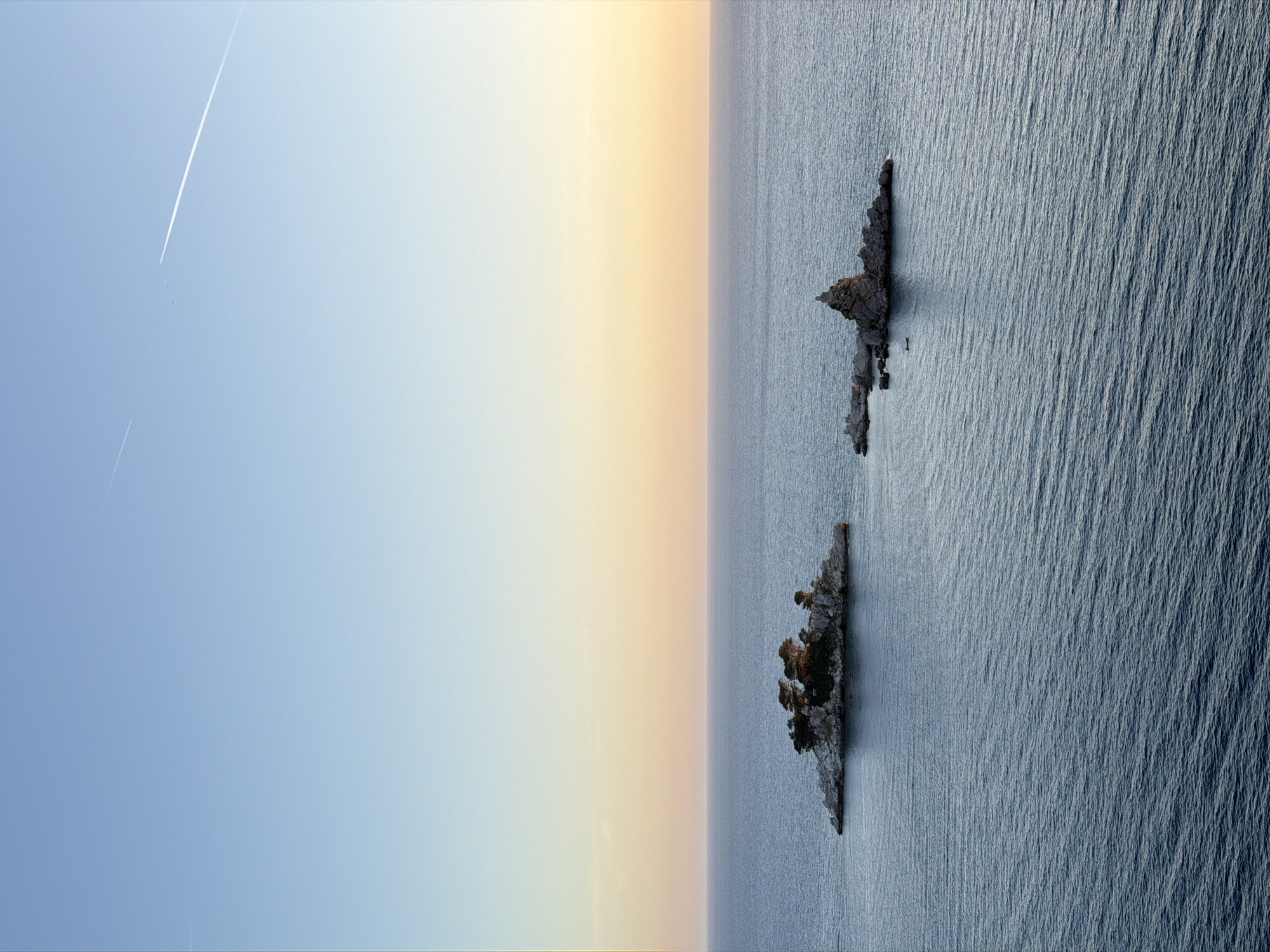 The image depicts a serene coastal landscape near Petrovac na Moru, Montenegro, at either sunrise or sunset. The sky presents a subtle gradient, transitioning from a light blue in the upper segment to a soft yellowish-orange hue closer to the horizon. Two faint white contrails are visible in the upper sky, one extending diagonally from the upper right and another shorter, thinner line in the upper left. The horizon line is sharply defined, separating the sky from a vast expanse of calm, dark blue-grey sea, characterized by subtle surface ripples.
Two distinct rocky islets emerge from the water. The larger islet on the left, identified as Katic, is densely covered with numerous dark green coniferous trees, appearing to be pines or cypresses, rooted amidst its rocky structure. To its right, the smaller islet, identified as Sveta Nedjelja, consists primarily of jagged rock formations, with a prominent, spire-like peak on its eastern side and minimal to no visible vegetation. A small, dark-colored boat is positioned in the water between the two islets, slightly closer to the Sveta Nedjelja islet. There are no individuals or explicit actions depicted.