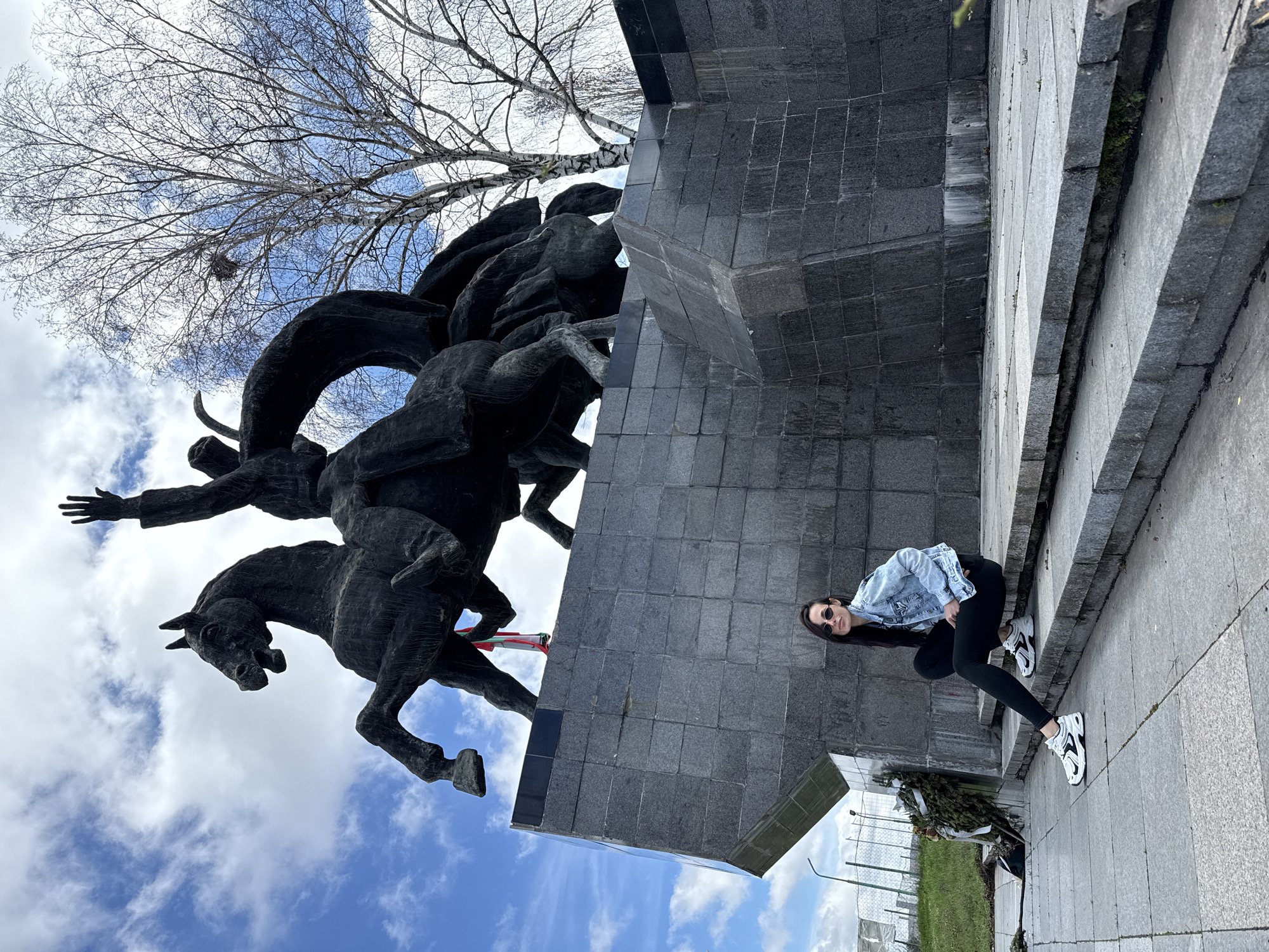 The image depicts the Monument to the Liberators in Pleven, Bulgaria. This large, dark-colored equestrian statue, likely bronze, features a central figure on a rearing horse with its right arm raised, accompanied by additional figures and horses. A red, green, and white ribbon is tied to the left front leg of the main horse. The monument rests on a substantial, multi-faceted pedestal constructed from dark grey granite blocks. At the base of the pedestal, to the left, are several withered wreaths. A female individual with long dark hair, wearing dark sunglasses, a light blue denim jacket, a dark top, black leggings, and white sneakers, is seated on the lower granite steps of the monument's base, facing the camera. The sky is partly cloudy with significant blue patches. To the right of the monument, a large, bare-branched deciduous tree, possibly a birch, stands with a bird's nest visible in its upper branches. To the left, a grassy incline leads to a wire fence, beyond which distant structures are visible under a bright sky. The foreground consists of grey paving stones forming steps, with minor vegetative growth in their crevices.