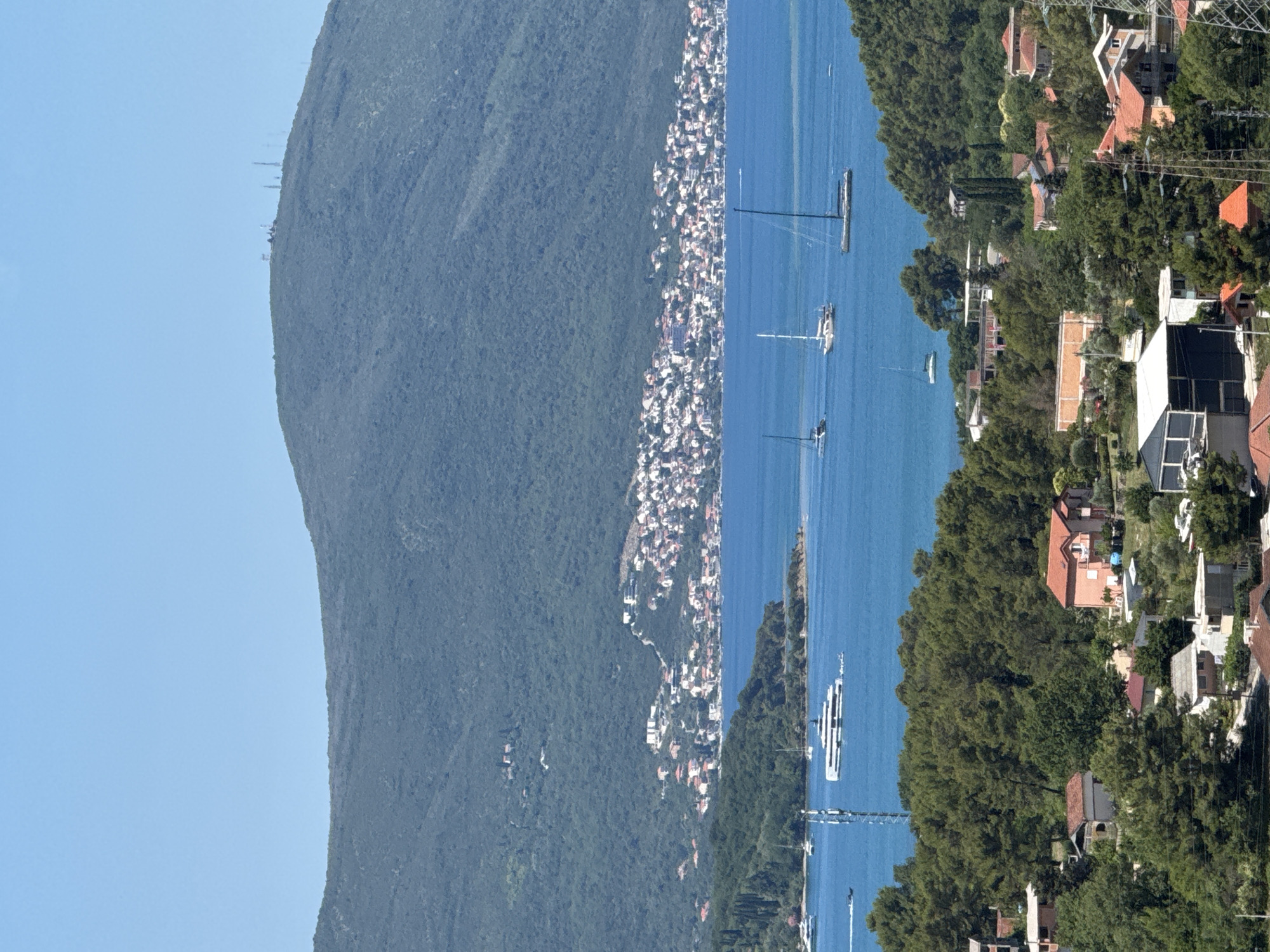 The image captures a panoramic view of a coastal landscape, likely in Kavač, Montenegro, on a clear day with a bright blue sky. The foreground features a dense expanse of green trees and several residential buildings with terracotta roofs, nestled on a hillside. These structures vary in size and architectural style, suggesting a mix of older and more modern dwellings.
Further back, a serene body of water, possibly a bay or gulf, is visible. Several boats, including sailboats and a large white yacht, are scattered across the water, indicating recreational activity. Along the shoreline of the water, a town or village with numerous white and red-roofed houses is densely built on the slopes of a large, tree-covered mountain that dominates the background. The mountain itself has a rugged, natural appearance, with some telecommunication towers sparsely visible on its summit.
The overall scene depicts a tranquil coastal environment where nature and human habitation coexist, characterized by lush greenery, clear waters, and a picturesque settlement against the backdrop of a majestic mountain.