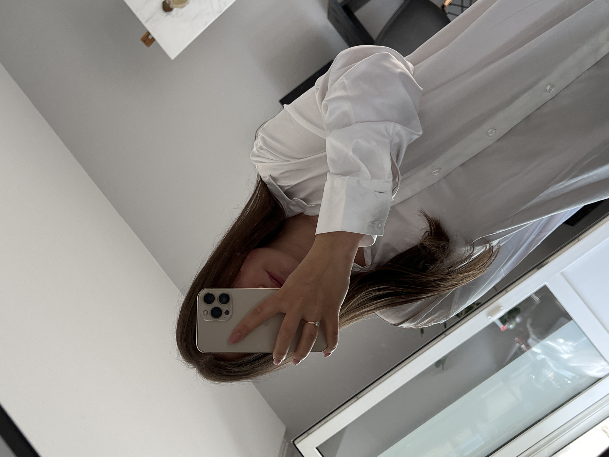 A woman is taking a selfie. She is wearing a white button-down shirt and holding a smartphone. The background includes a table, a chair, a wall, and a window. It is not possible to determine the city of Ternopil, Ukraine, from this image.