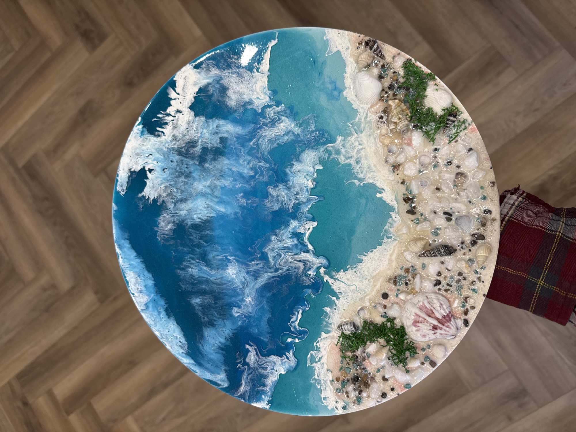 The image displays an overhead view of a circular resin art piece, likely a tabletop, that vividly depicts an ocean and beach scene. The left and central sections of the circle are filled with the "ocean," rendered in various shades of blue, ranging from deep azure to lighter turquoise. White, swirling patterns, characteristic of resin art, create the illusion of frothy waves and seafoam, conveying a sense of movement and depth.
The "ocean" seamlessly transitions into a textured "sandy beach" area on the right side of the circle. This beach section is a light, sandy color and is richly embellished with an assortment of embedded natural elements. These include numerous seashells of different types, such as conch shells, clam-like shells, and smaller spiral shells. Interspersed among the shells are small, smooth pebbles and stones in shades of black, white, and greenish-blue. Patches of bright green, moss-like material are also visible, adding a touch of simulated vegetation to the beach.
The entire artwork has a glossy finish, indicative of resin. The background features a warm-toned wooden floor with a distinct herringbone pattern. A small portion of a red and plaid fabric, possibly a sleeve or blanket, is visible on the right edge of the frame. There is no discernible activity taking place, and no text is visible in the image.