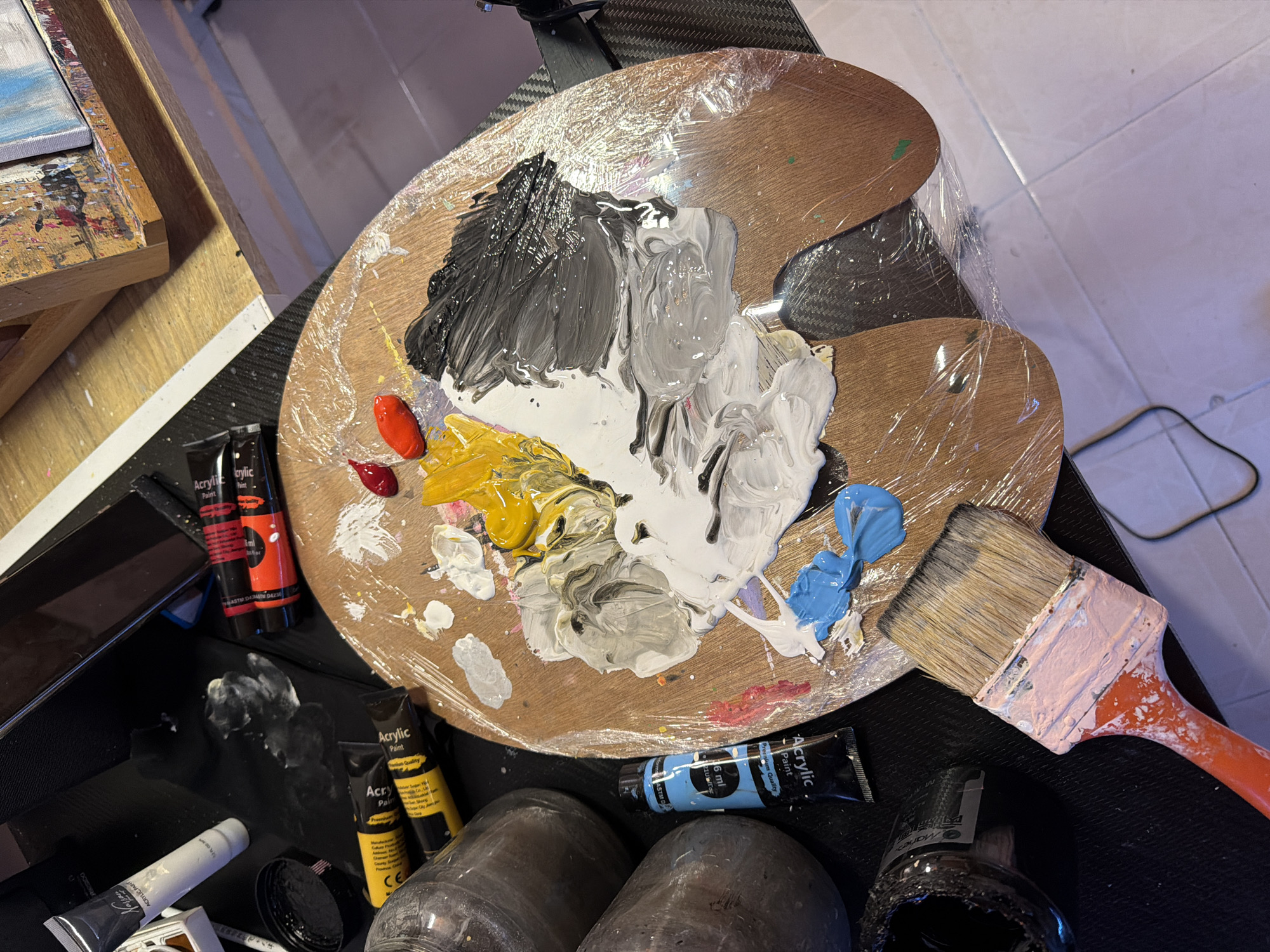 The image depicts an art studio environment. The foreground features a wooden artist's palette, covered in plastic wrap, with various colors of acrylic paint swirled together, including white, grey, black, yellow, red, and blue. Two tubes of acrylic paint, one blue and one white, are lying near the palette, as well as three smaller tubes of red and orange paint. To the left of the palette are two large glass jars, one open and containing a dark substance. A paintbrush with an orange handle and light pink ferrule, coated with pink paint, rests in the lower left corner of the frame. In the background, part of an easel with a canvas is visible, and other art supplies are scattered around. The scene appears to be an indoor workspace, with a tiled floor visible in the lower right corner. There are no people or specific identifiable landmarks of Svilengrad, Bulgaria.