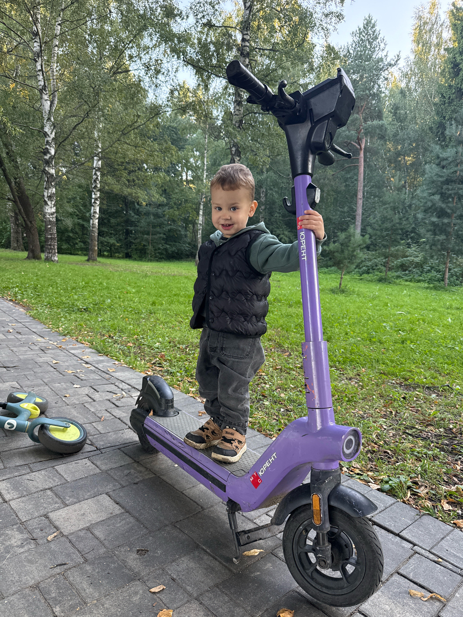 A young boy, approximately two years old, stands on a purple electric scooter on a paved pathway in a park. The boy is wearing a dark puffer vest over a light green hooded sweatshirt and dark gray pants. He has light brown hair and is looking towards the camera with a smile. His right hand is gripping the scooter's handlebar. The scooter is a rental scooter, indicated by the logo and branding on the steering column. To the left of the scooter, a smaller, green and yellow child's balance bike lies on its side on the pathway. The pathway is made of gray interlocking bricks. Beyond the pathway is a grassy area with scattered fallen leaves. Behind the grass is a dense line of trees, including birch trees with white bark and evergreen trees. The sky is visible through the upper branches of the trees and appears to be overcast. The scene appears to be set in the city of Rusanovo, Russia, given the context provided.