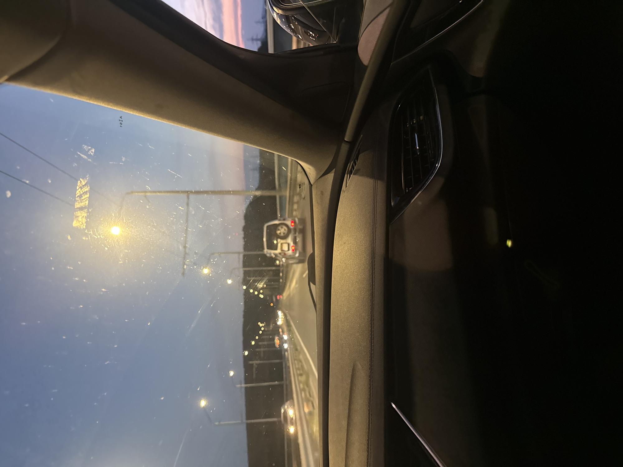 The image captures a forward view from the interior of a vehicle, driving on an elevated roadway during twilight hours in Varna, Bulgaria. The vehicle's dark-toned dashboard, featuring stitching details and a chrome-accented air vent on the right, occupies the lower foreground. A dark, fabric-covered A-pillar frames the view on the right. A small, illuminated green indicator light is visible on the lower dashboard area. In the upper right quadrant of the windshield, the inverted text "rEjA" is present.
The windshield is notably soiled with numerous specks and streaks, causing external light sources to appear diffused and fragmented. Ahead, a white SUV with a spare tire mounted on its rear is visible in the same lane. Multiple other vehicles with illuminated headlights are observed further down the multi-lane road. The roadway is lined with numerous streetlights on poles, emitting bright yellow light that reflects off the road surface. The sky transitions from a deep blue-grey overhead to lighter tones with hints of pink and orange on the right horizon. Distant forested hills are silhouetted against the sky on the left, while utility poles or similar structures are faintly visible against the horizon on the far right. The right side mirror of the vehicle reflects ambient lights from the side or rear.